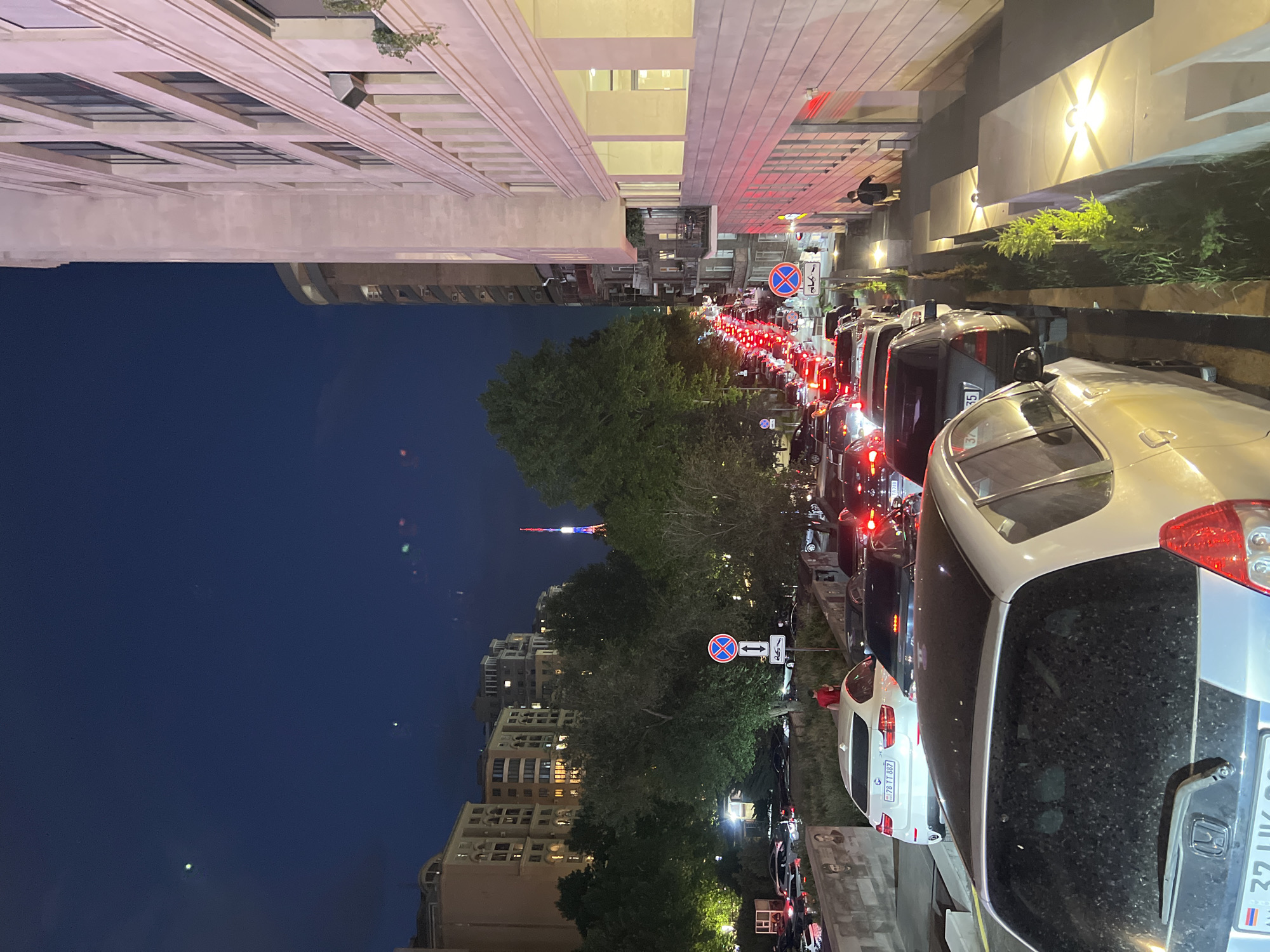 The image captures a nighttime urban street scene, likely in Yerevan, Armenia. The dark blue sky indicates late evening or night, with artificial lights illuminating the surroundings.
On the right side of the frame, numerous cars are parked along the curb, with several more vehicles visible in a long line extending down the street, suggesting a traffic jam. Their red taillights glow brightly, creating streaks of light in the distance. The closest parked vehicle is a silver SUV with an Armenian license plate partially visible as "33 UK 08". Further down, a white BMW also shows a partial Armenian license plate "78 TT 867". A person wearing a red top is faintly visible among the parked cars.
To the left, a tall, modern building with a light-colored facade and prominent horizontal architectural features rises high, its windows reflecting some light. Below this building, a line of green trees separates the foreground from the background. Through and above the trees, several multi-story residential buildings with illuminated windows are visible. In the far distance, a slender, brightly lit tower, resembling the Yerevan TV Tower, stands out against the dark sky.
Notable details include several traffic signs: a clear "No Stopping" sign (a red circle with a blue background and a diagonal red line) and a rectangular blue sign with white arrows beneath it. The overall scene conveys the atmosphere of a busy city street after dark.