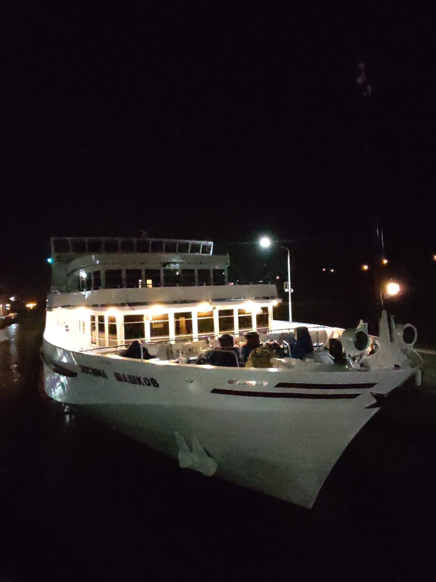 The image depicts a white passenger vessel, likely a river cruise ship, at night in an unspecified location of the city of Unknown, Unknown. The vessel has multiple decks with illuminated windows, suggesting an active or recently active operational status. Passengers are visible on the main deck, seated in what appear to be outdoor seating arrangements. The ship's name, "ZOCHMA MAHIKOB," is discernible on its hull. The surrounding environment appears dark with minimal background light sources, possibly indicating a nighttime river or dock setting.