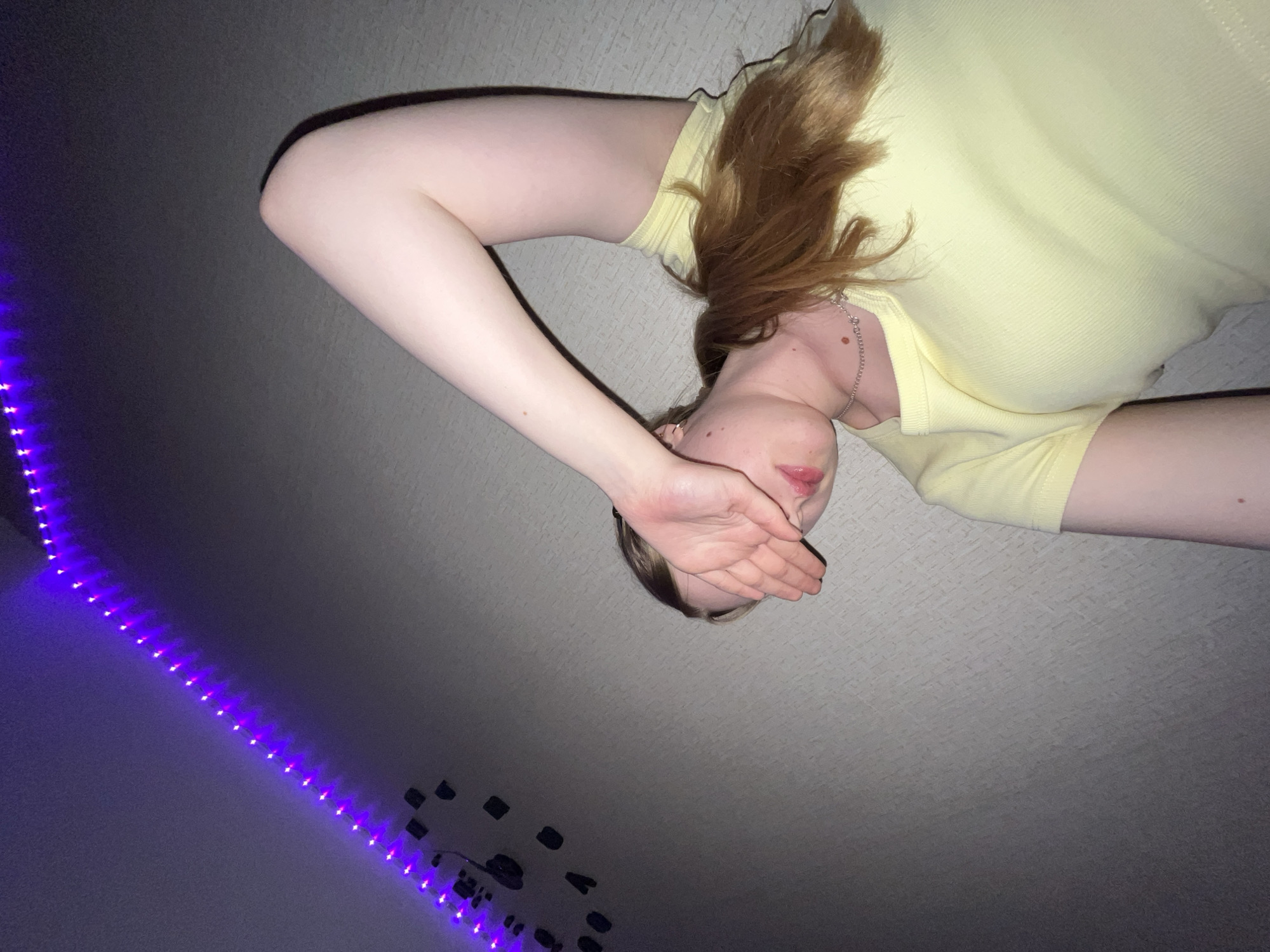 A young person is lying on a carpeted floor, possibly in a room with purple LED strip lights on the wall. They are wearing a light yellow top and have reddish-blonde hair. Their right hand is raised to cover their face. The scene is upside down, taken from a low-angle perspective. There are black adhesive squares visible on the wall, near the LED strip lights. The location is not discernible from the image and therefore the claim of being in the city of Chaikovsky, Russia cannot be verified.