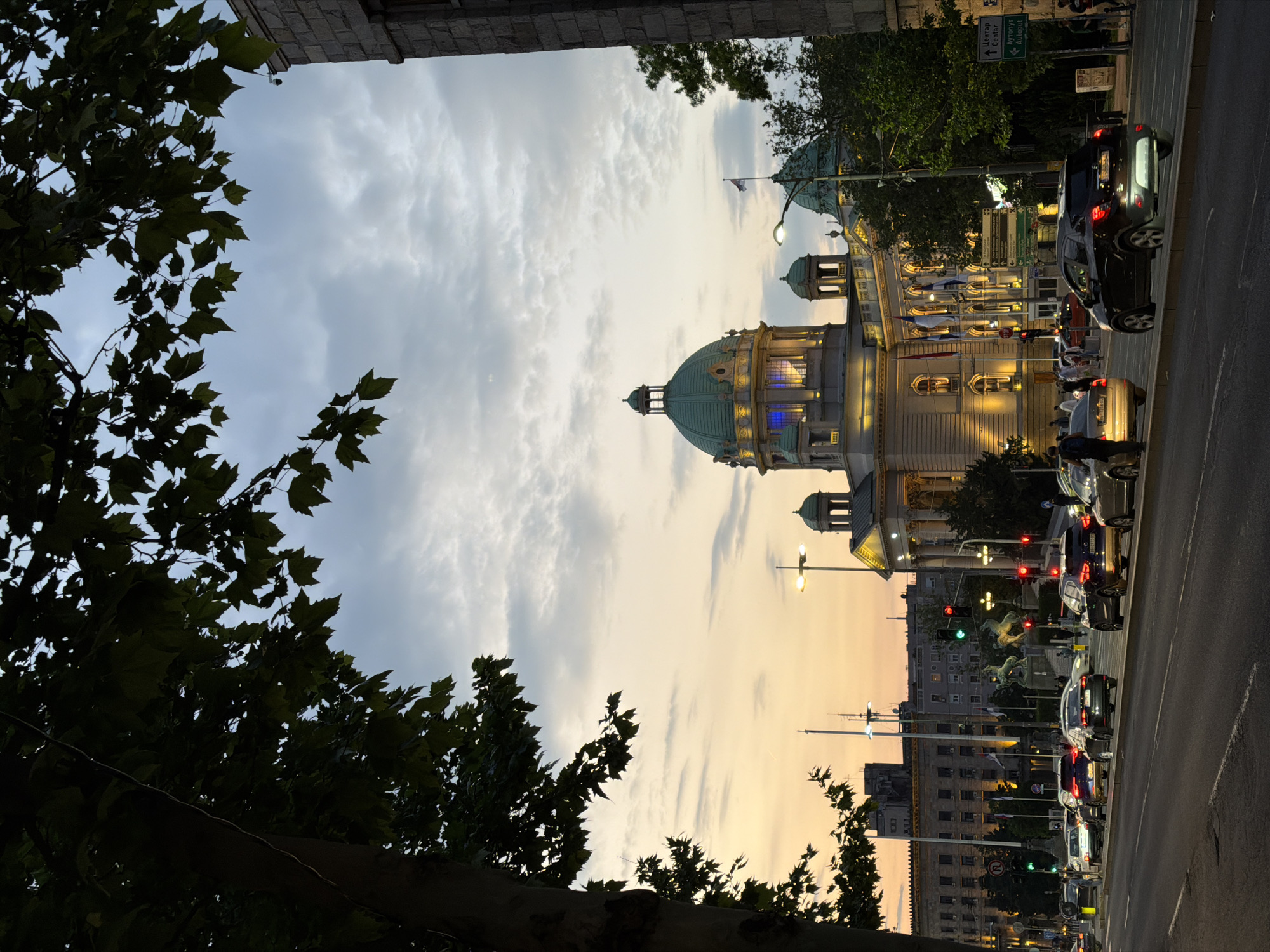 The image shows the Church of Saint Sava, a Serbian Orthodox church located on the Vračar plateau in Belgrade, Serbia. The building's dome is visible against a backdrop of a cloudy sky. The street beside the church is lined with parked cars and street lamps. Trees and buildings frame the image. The scene appears to be at dusk, with warm lighting illuminating the church.
