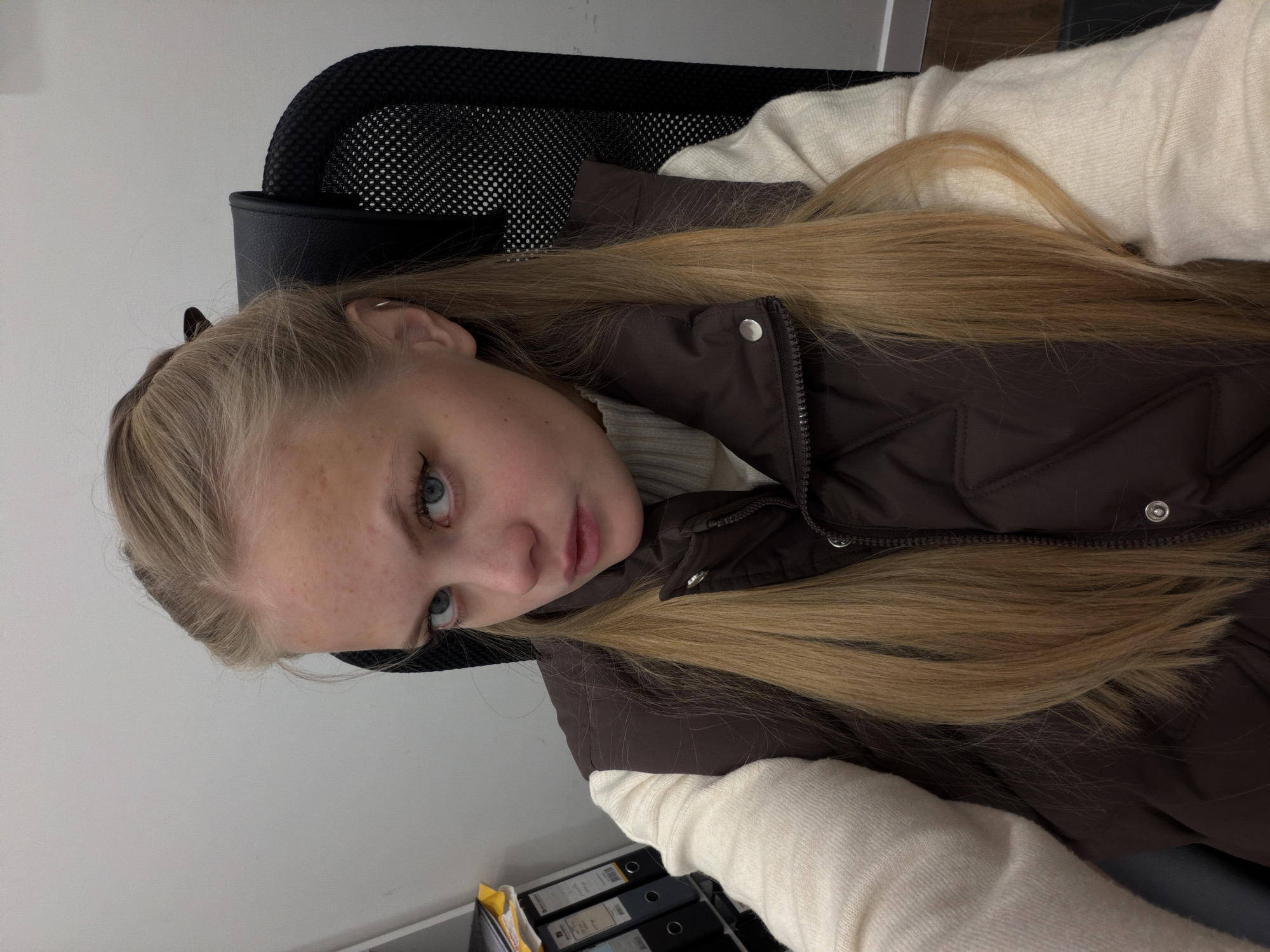 A young fair-skinned person with long blonde hair is seated in a black mesh office chair. The person is wearing a white sweater and a brown puffer vest. The background consists of a white wall, a black chair, and a shelf with binders on it. There is no evidence to suggest the image was taken in Izhevsk, Russia.