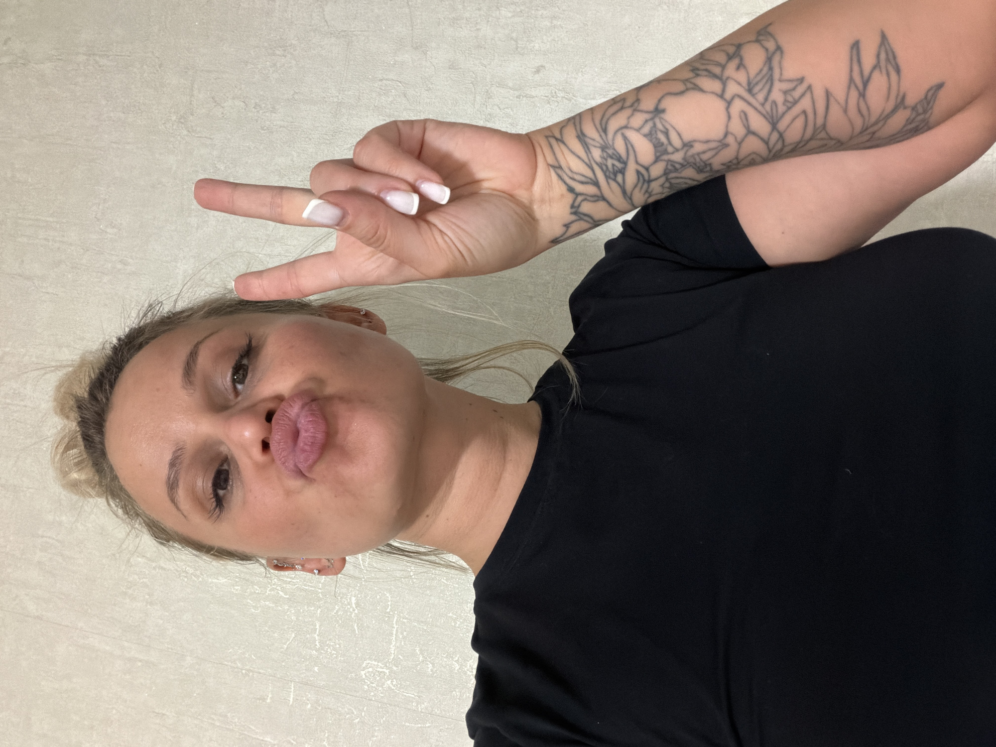 A fair-skinned woman with blonde hair pulled back in a bun poses for a selfie. She is puckering her lips and making a peace sign with her right hand. She is wearing a black t-shirt. Her left arm, adorned with a floral tattoo, is visible. The background is a neutral, textured wall. There is no indication of the location being in Sarapul, Russia.