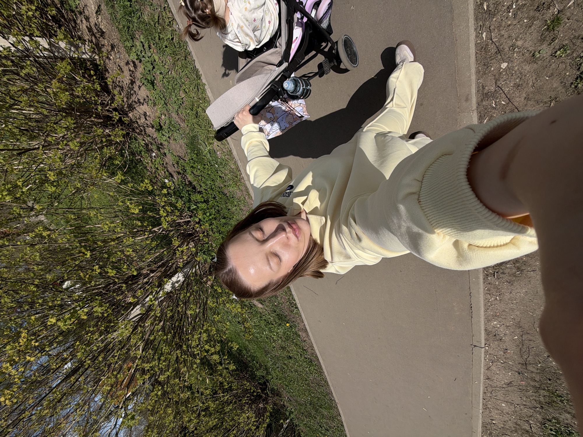 Captured from a high-angle perspective, the image presents a woman dressed in a light-yellow sweat suit, seemingly taking a selfie on a paved walkway. To her left, a stroller is visible, occupied by a child. The walkway is bordered by greenery on either side. The location is Reshetnikovo, Russia.