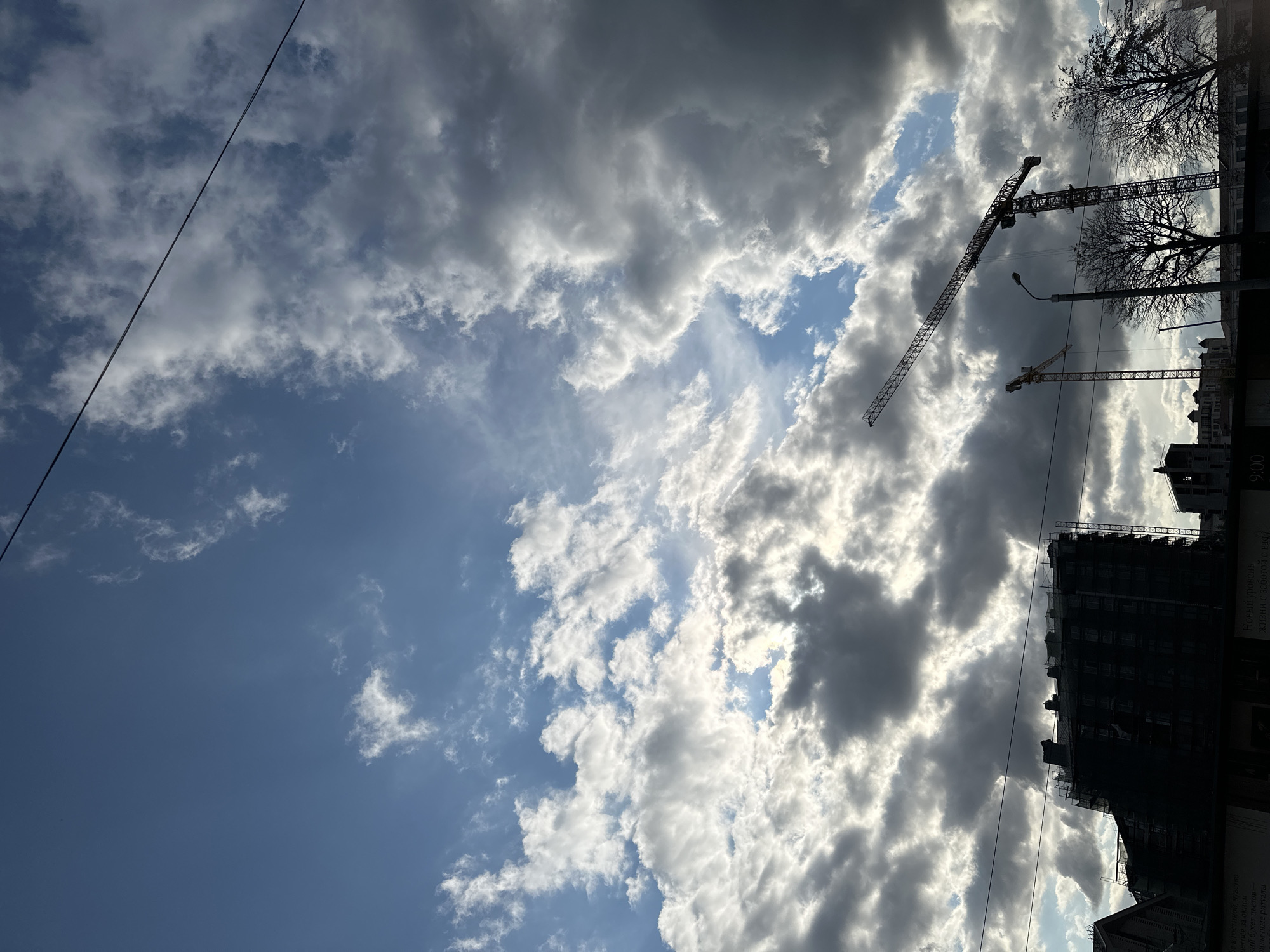 This is a low-angle, medium shot of a cloudy sky above a construction site in Minsk, Belarus. Two construction cranes are visible against the sky, one tall and thin, the other shorter and wider. In the lower portion of the frame, the silhouette of a multi-story building under construction is visible, covered in scaffolding and netting. Bare-branched trees are also visible in the foreground, partially obscuring the construction site. A single power line stretches diagonally across the upper portion of the image. The sky is filled with dramatic, partially sunlit clouds, suggesting a dynamic weather system. The overall scene conveys a sense of ongoing urban development and industrial activity.