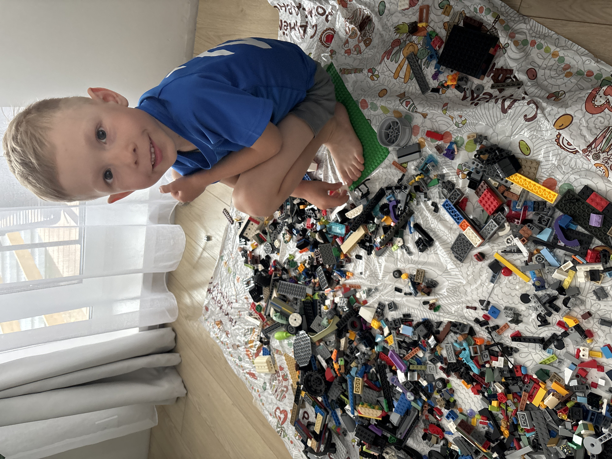 A child with blond hair, wearing a blue t-shirt and grey shorts, sits cross-legged amidst a large pile of Lego bricks. The bricks are scattered on a patterned sheet spread out on a wooden floor. A window with white blinds is visible in the background. The location is in a room in an apartment building in the city of Chaikovsky, Russia.