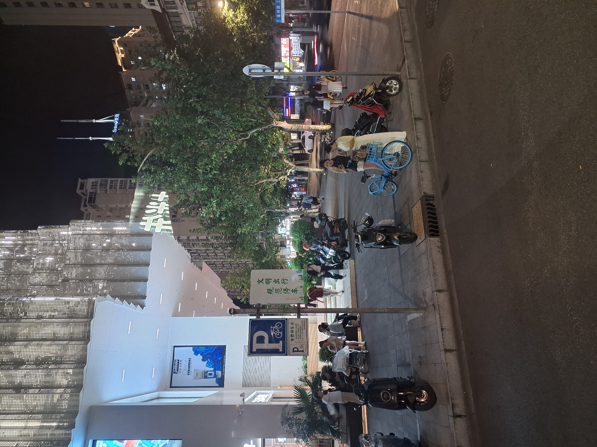 The image captures a street scene at night in Shanghai, China. The foreground shows a sidewalk with several parked electric scooters and bicycles, some of which appear to be shared rental bikes. A few people are seated on a low wall along the sidewalk, including a woman holding a baby. A stroller is also present.
Further down the sidewalk, more people are walking or standing. A large building with a modern, illuminated facade dominates the background, its design featuring intricate latticework. Across the street, other buildings are visible, some with lit windows, suggesting commercial or residential activity. Streetlights illuminate the scene, casting long shadows and highlighting the urban environment. A street sign with Chinese characters indicates the location, and a blue traffic sign with a 'P' and a bicycle symbol denotes a parking area. The overall atmosphere is that of a bustling urban thoroughfare at night, with people and vehicles interacting within the cityscape.