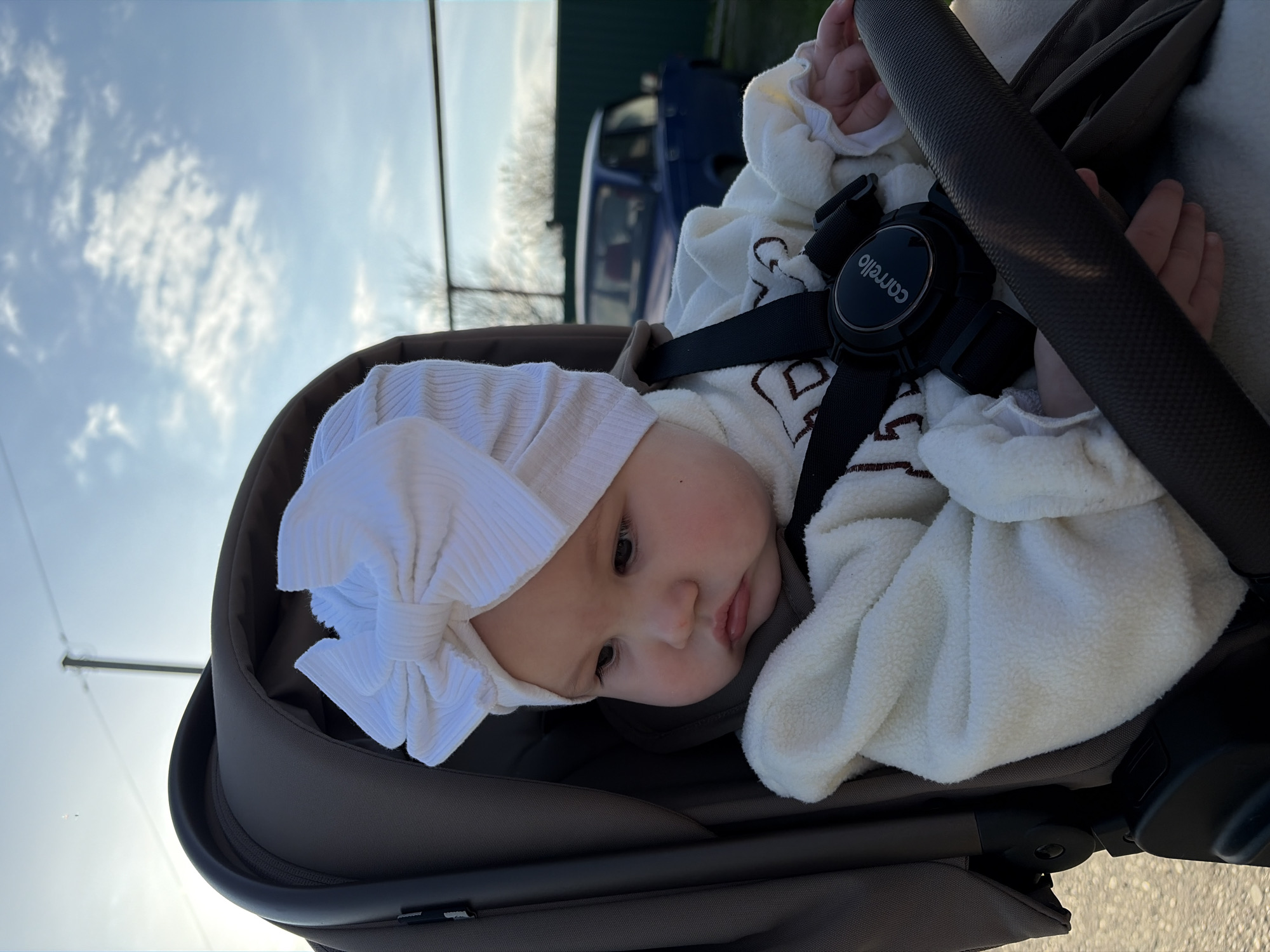 The image, originally rotated 90 degrees counter-clockwise, portrays a baby resting in a stroller or car seat during an outdoor excursion in Novopokrovskaya, Russia. The baby is the central subject, viewed from a slightly elevated perspective. It wears a white, ribbed headwrap with a large bow and a cream-colored, fluffy garment, possibly fleece, over a lighter top. The baby's eyes are open, and its expression is neutral, with a subtle pout, suggesting calm or contemplation.
The baby is secured in the seat by a black harness system. The central buckle of this harness clearly displays the brand name "Carrello". The stroller itself appears to be a dark brown or grey color.
The background reveals an outdoor setting under a bright, partly cloudy sky. In the mid-ground, a blurred blue structure, possibly a vehicle or a building, is visible, along with faint outlines of bare trees. The ground immediately surrounding the stroller is indistinct but appears to be a paved or concrete surface. The lighting suggests it is daytime, with soft, diffused sunlight, likely morning or late afternoon.