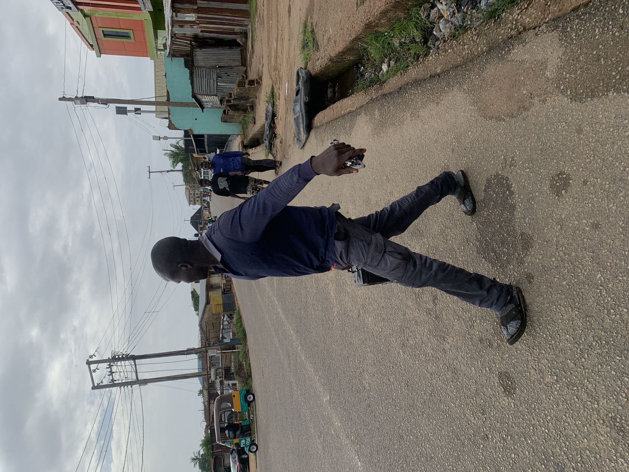 A man is walking down a street in Jos, Nigeria. The street is paved and has a drainage ditch on the right side. There are buildings on both sides of the street, including some with corrugated metal walls. There are other people walking in the distance. A few auto rickshaws are parked on the side of the road. Electrical poles and wires are visible overhead. The sky is cloudy.