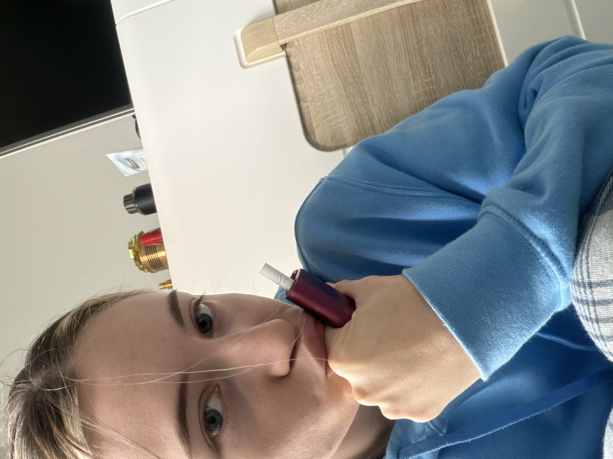 A close-up, angled selfie captures a young person, likely female, holding a reddish-purple electronic device to her lips. The device has a white stick, resembling a cigarette, inserted into its top. Her gaze is directed towards the camera, with a neutral expression. Loose strands of light brown hair cross her face, particularly prominent across her nose and cheeks. She is wearing a light blue hooded sweatshirt, visible on her arms and torso. To her right, a pattern that appears to be plaid or checkered can be seen on another piece of clothing.
The background is a mix of indoor elements. To the left side of the image (top if the image were re-oriented), a white wall or piece of furniture dominates. Below it, a dark screen or monitor is partially visible, followed by a small, white packet with blue markings, possibly a personal care item. Further down, a cylindrical object with a metallic, possibly brass or gold-toned, cap and red accents is visible, resembling a pipe fitting or part of a fixture. To the right (bottom if re-oriented), a wooden surface with a light grain, possibly a desk or the arm of a chair, is present against another white surface. The lighting is bright and even, suggesting an indoor setting during the daytime. No clearly decipherable text is visible on the objects or in the background, although there are faint markings on the white stick in the device and on the small packet.