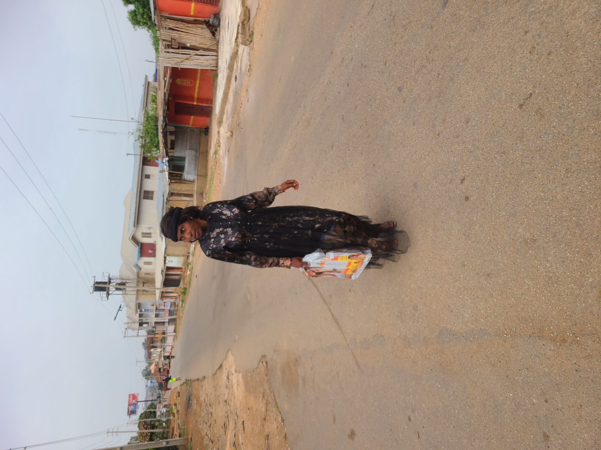 A woman in a black floral dress and head covering walks along a paved road in Jos, Nigeria. She carries a plastic bag. Buildings and utility poles line the side of the road.