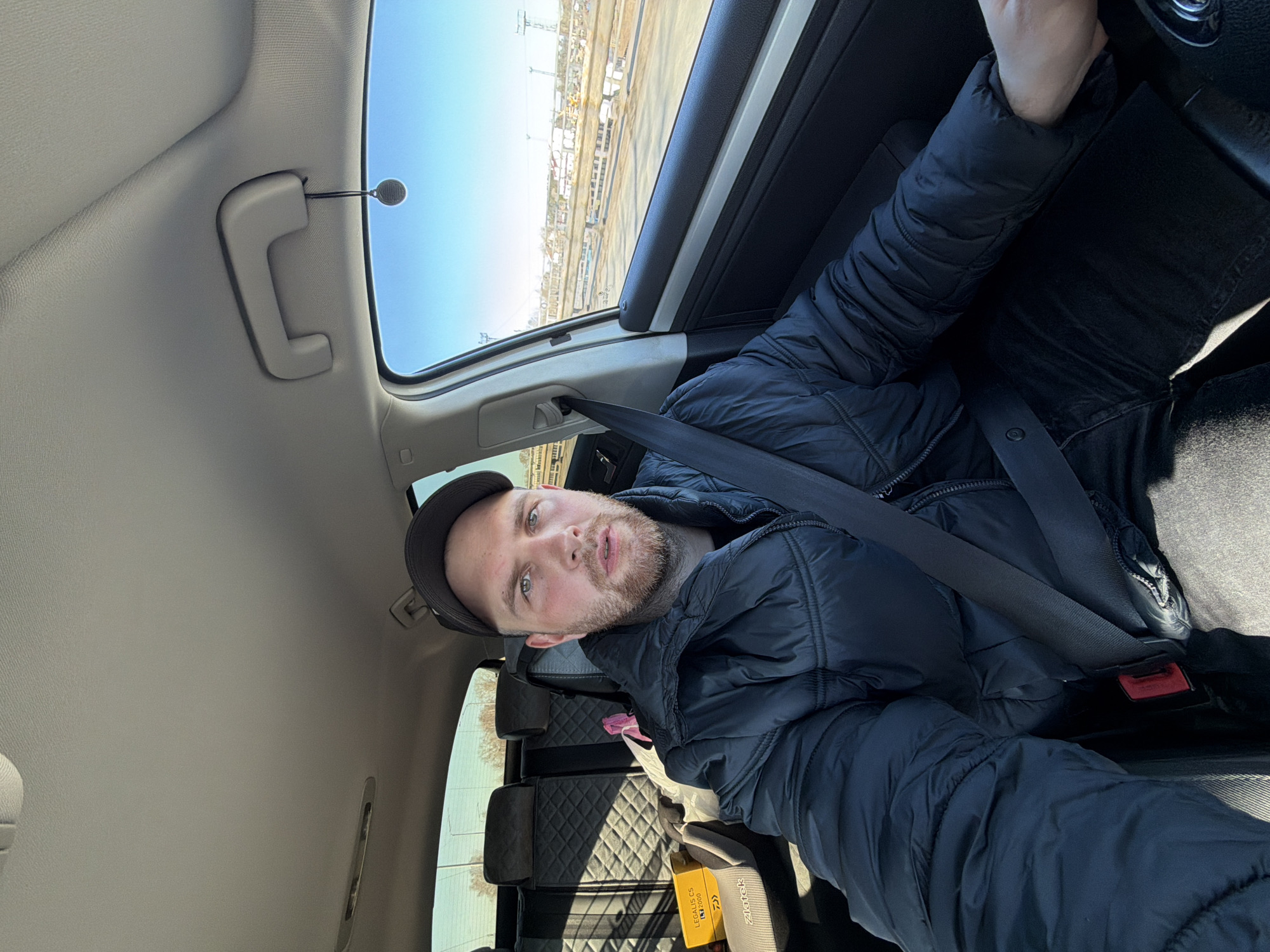 A man with light skin, short hair, and a beard is in the driver's seat of a car. He is wearing a dark blue jacket and a cap, and the seatbelt is fastened. The car is likely moving, as evidenced by the blurred landscape visible through the sunroof. The landscape appears to have some kind of industrial construction elements. In the back seat, there are personal belongings, including what appears to be a package labeled "Legalitas CS 2000" and "Ziatek". Based on the available information and the label, the location may be somewhere around the city of Chaikovsky, Russia.