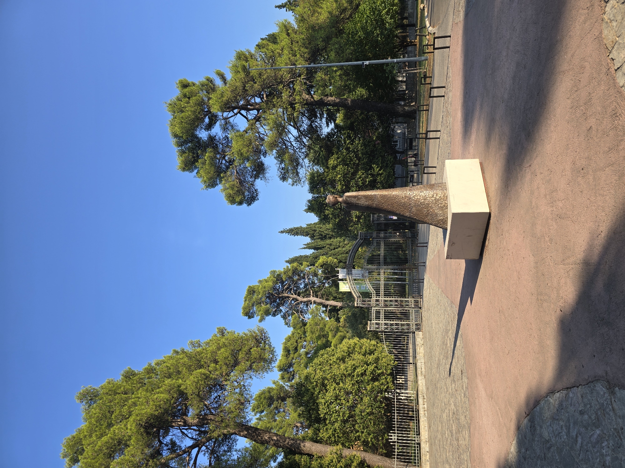 The image depicts a scene in Podgorica, Montenegro, featuring a bronze sculpture of a figure in a flowing robe, standing on a light-colored pedestal. The sculpture is positioned in the foreground, casting a long shadow on a pink paved walkway. Behind the sculpture, a decorative metal gate leads to a park-like area with lush green trees and foliage. In the background, a clear blue sky dominates the upper portion of the image. The overall atmosphere is serene and bathed in natural light, suggesting it was taken during daytime.