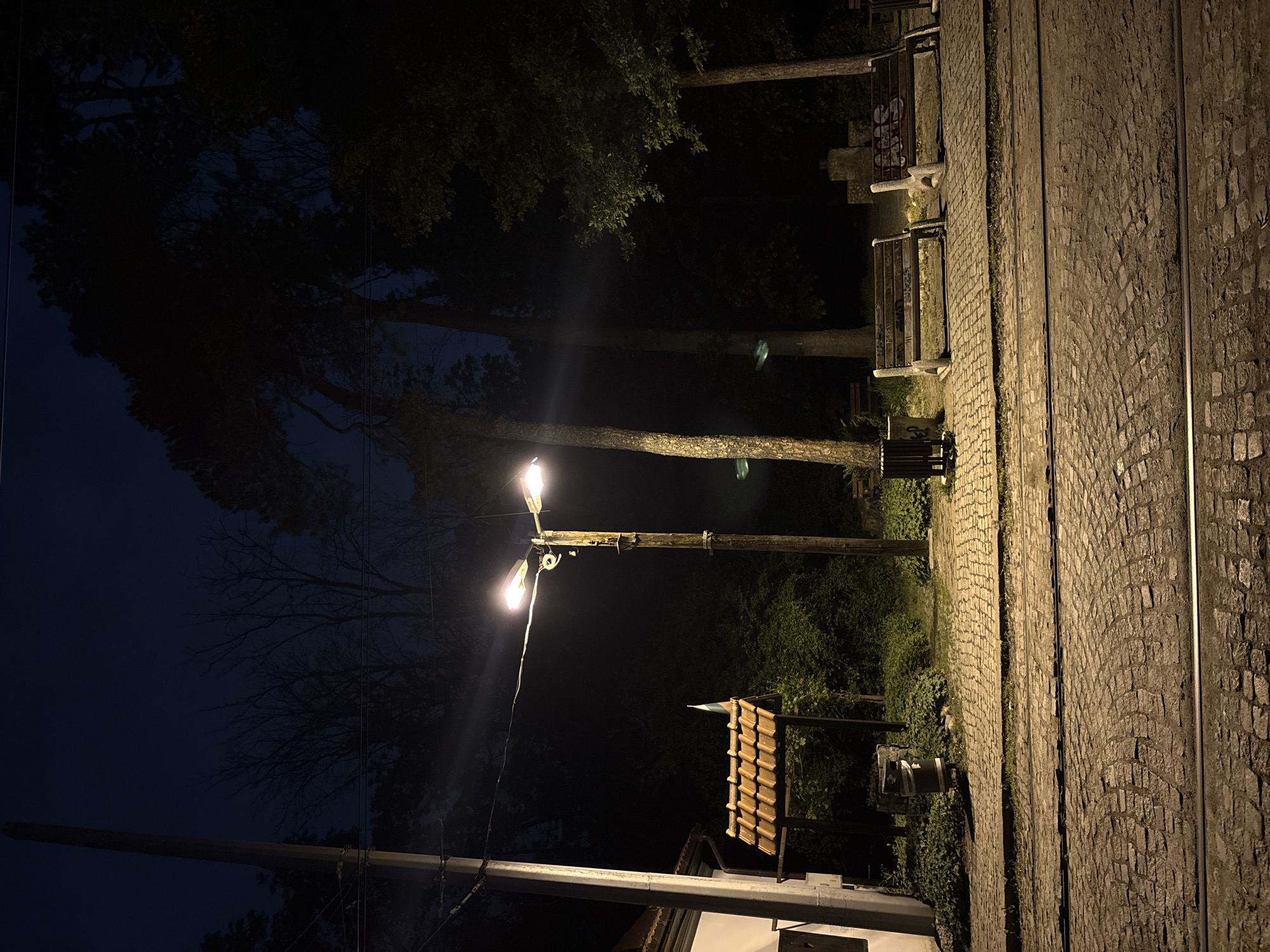 This nighttime scene shows a section of pavement in Sofia, Bulgaria, possibly near a tram line. The pavement is constructed from cobblestones. Parallel rails run along the edge of the pavement. Benches and a trash bin are located along the edge of the walkway. Further back, there is a small tiled structure with a roof. A lamppost illuminates the space with two bright lights. Trees and other vegetation are visible in the background. The sky is a dark blue. The bottom of a white building is visible in the lower portion of the frame.