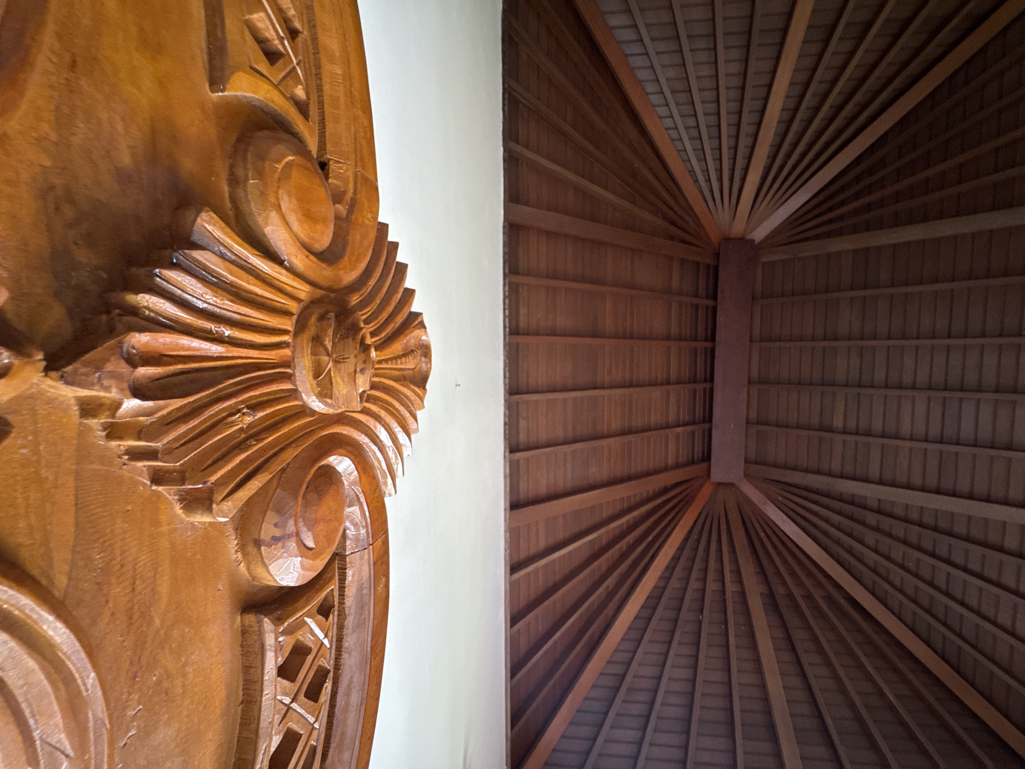 The image shows a detail of a carved wooden structure next to a ceiling with a radial pattern of wooden slats and beams. The carved structure is on the left side of the image and exhibits intricate designs and a light brown color. The ceiling occupies the right side, and its wooden elements converge towards a central point, giving a fan-like appearance. A plain white wall separates the carved wooden structure from the ceiling. The image was taken in Kuta Selatan, Indonesia.