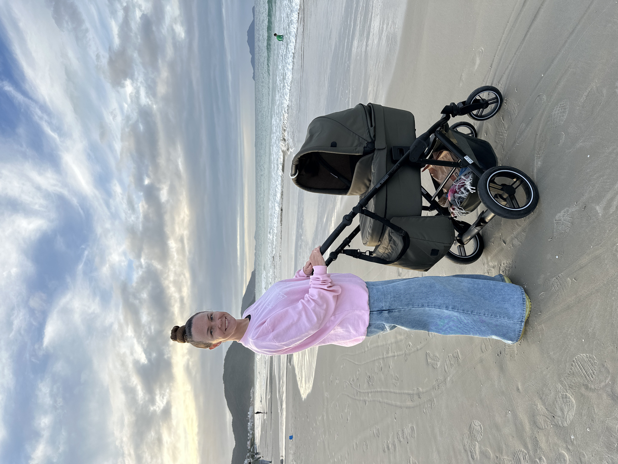 A woman stands on a beach next to a baby stroller. The beach appears to be located in Florianópolis, Brazil, judging by the landscape. The woman is smiling at the camera, she is wearing a pink sweatshirt and blue jeans. The stroller is dark green and has a baby inside. The sky is cloudy. The ocean is visible behind the woman. There are mountains in the background.