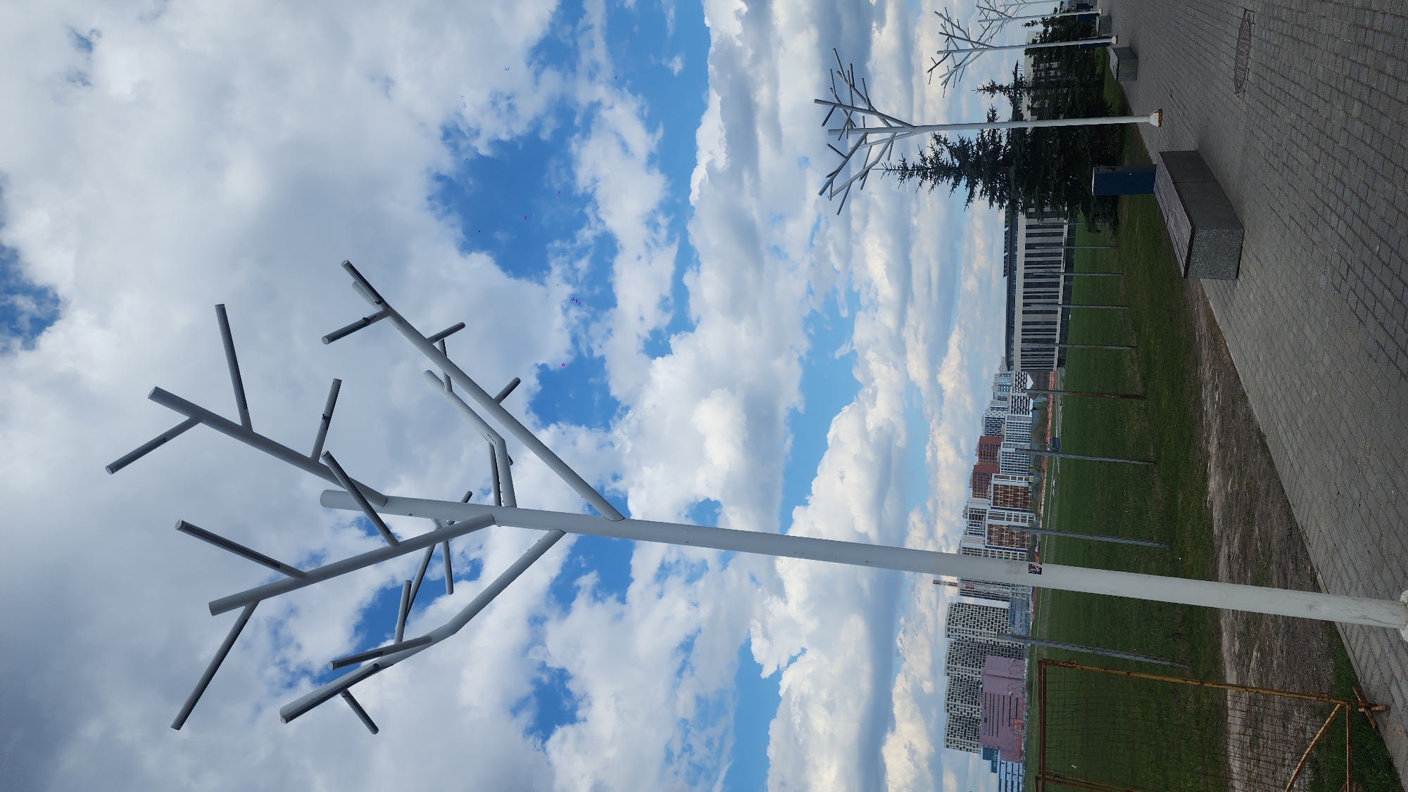 The image shows an outdoor scene in Moscow, Russia. A row of stylized, white, metallic "tree" sculptures stand on a green field alongside a paved walkway. In the background, there are apartment buildings and a larger institutional building. The sky is filled with clouds. A bench and a small evergreen tree are visible further down the path. A soccer goal is visible in the front right of the image.
