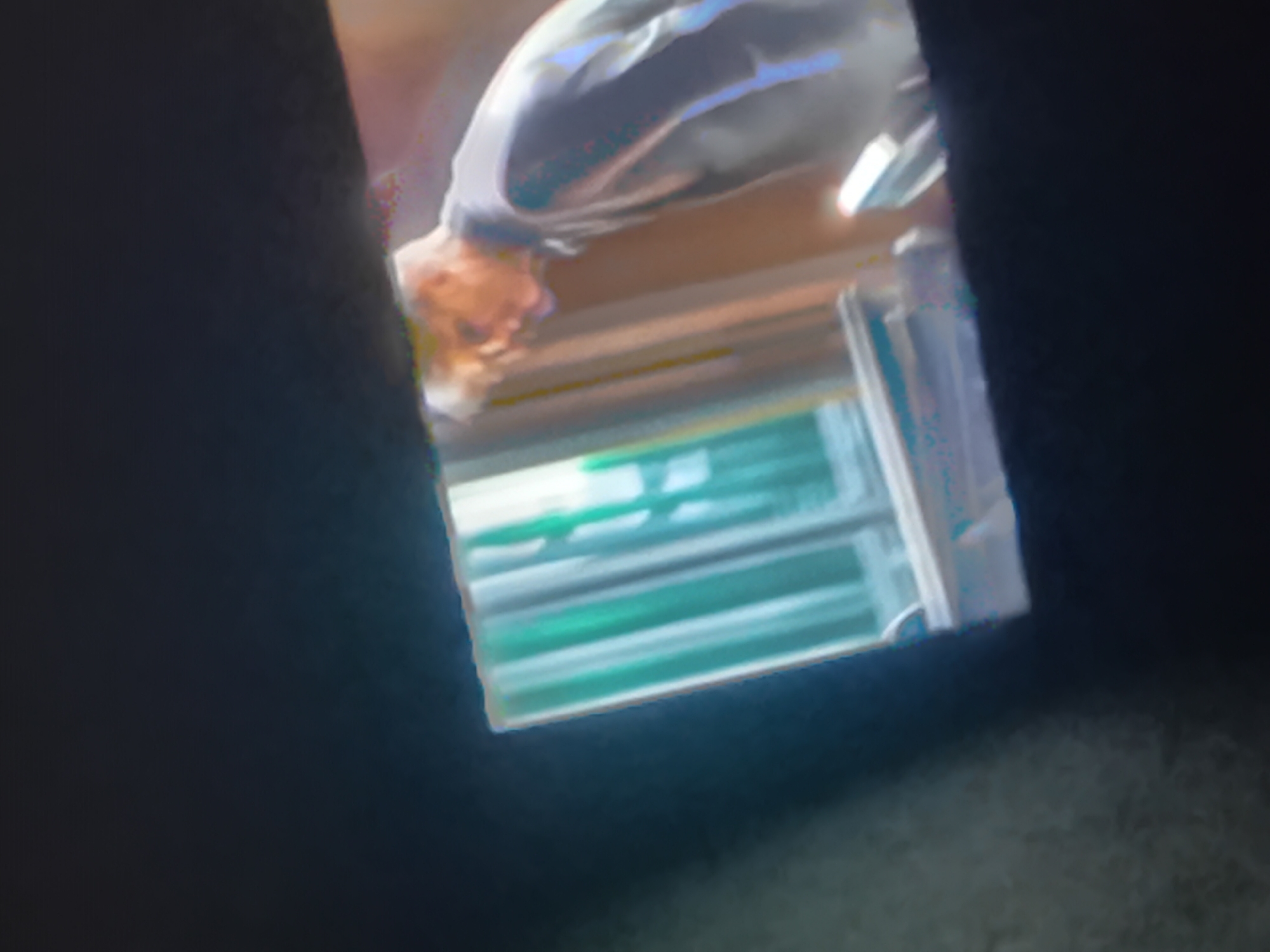 The media file shows a person bent over, their face visible in the center of the frame. The person has light-colored hair and is wearing a light-colored shirt. The background appears to be a blurred interior with horizontal lines, possibly indicating a window or blinds. The lighting is somewhat dim, but the face of the person is illuminated. The image is tilted and somewhat out of focus, suggesting it might be a candid shot or taken at an unusual angle. There is no discernible text in the image. The overall impression is of a casual or potentially private moment, with the focus on the person's posture and facial expression, though the emotion is hard to determine due to the blurriness. The surrounding darkness and the way the image is framed suggest it was taken within a confined space, possibly indoors.