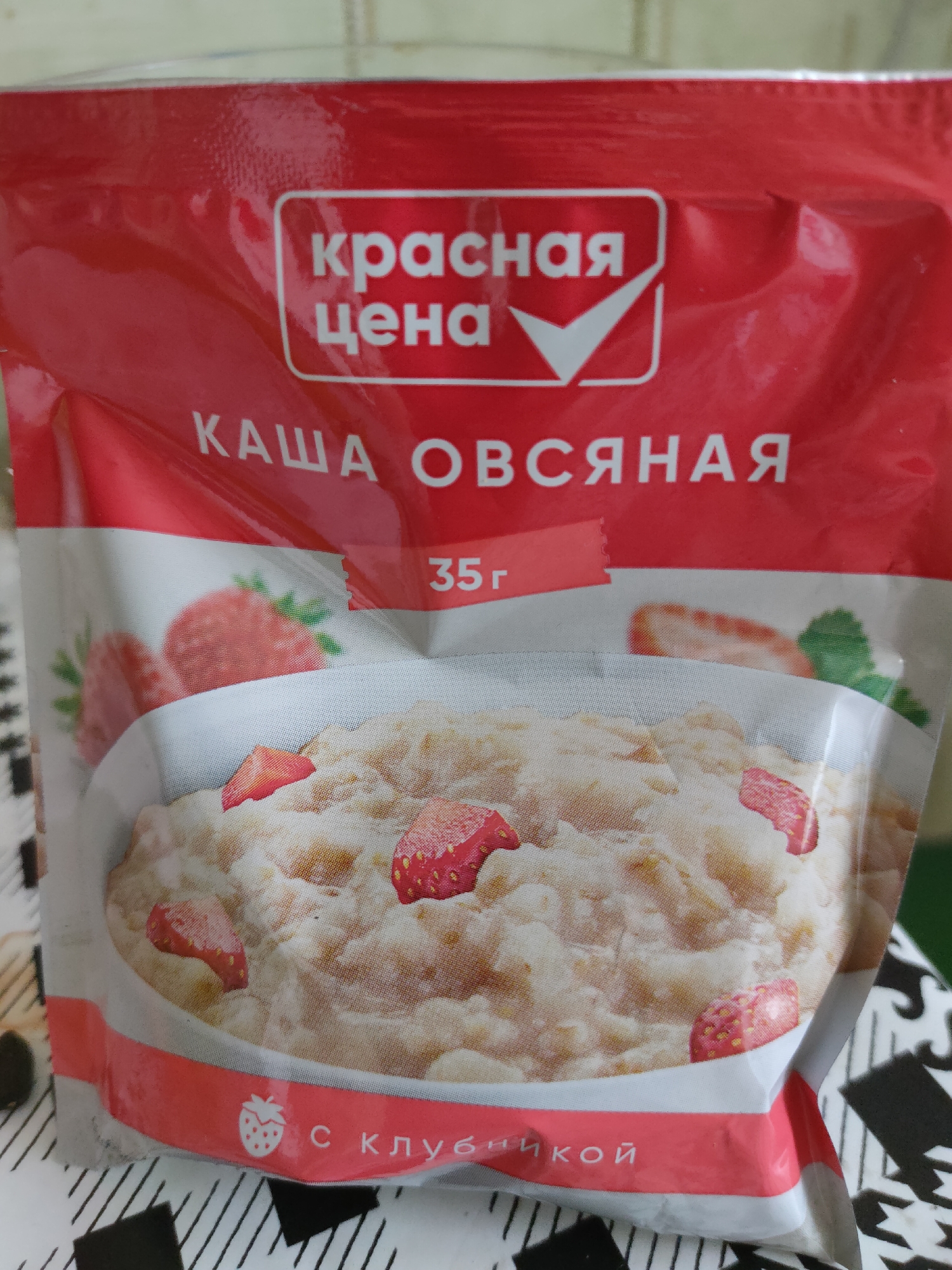 The image shows a close-up of a red package of oatmeal with strawberries. The text on the package is in Russian. The brand name "Красная цена" (Red Price) is visible at the top, along with a white checkmark symbol. Below that, the product name "КАША ОВСЯНАЯ" (Oatmeal Porridge) is written in large white letters. A small red rectangle indicates "35г" (35 grams). The lower part of the package features an illustration of a bowl of oatmeal with sliced strawberries and whole strawberries. At the very bottom, the text "С КЛУБНИКОЙ" (With Strawberries) is written, accompanied by a strawberry icon. The packaging is slightly crinkled. The background is blurred but appears to be a textured surface, possibly a tablecloth or a table. There is no indication of the location in Oktyabrsky, Russia.