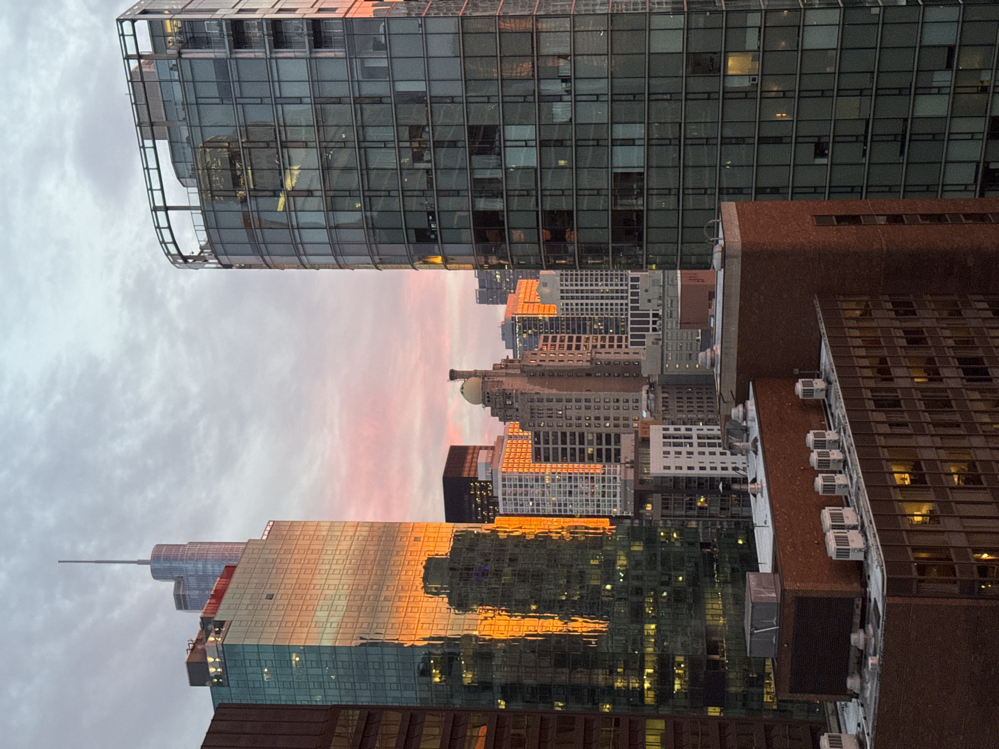 The image depicts a cityscape at dusk, likely Chicago, given the architectural style and density of the buildings. The sky is a mix of gray clouds and pastel pink and orange hues from the setting sun, casting a warm glow on the skyscrapers.
In the foreground, a lower-rise brick building with visible HVAC units on its roof occupies the bottom portion of the frame. To its right and behind it, several other buildings rise, with varying facade designs and lighting from interior offices.
Dominating the mid-ground and background are towering skyscrapers. On the left, a modern glass-facade building reflects the sunset in a vibrant orange hue. Adjacent to it, another skyscraper with a more segmented glass facade rises, with the distinct spire of the Willis Tower (formerly Sears Tower) visible in the distance. To the right, a curved glass skyscraper stands prominently, its numerous windows reflecting the dusky sky. Further back, a cluster of buildings with more traditional facades, including one with a domed roof, are visible.
The overall impression is one of urban density and the dramatic beauty of a city skyline at sunset. No people are visible in the image.