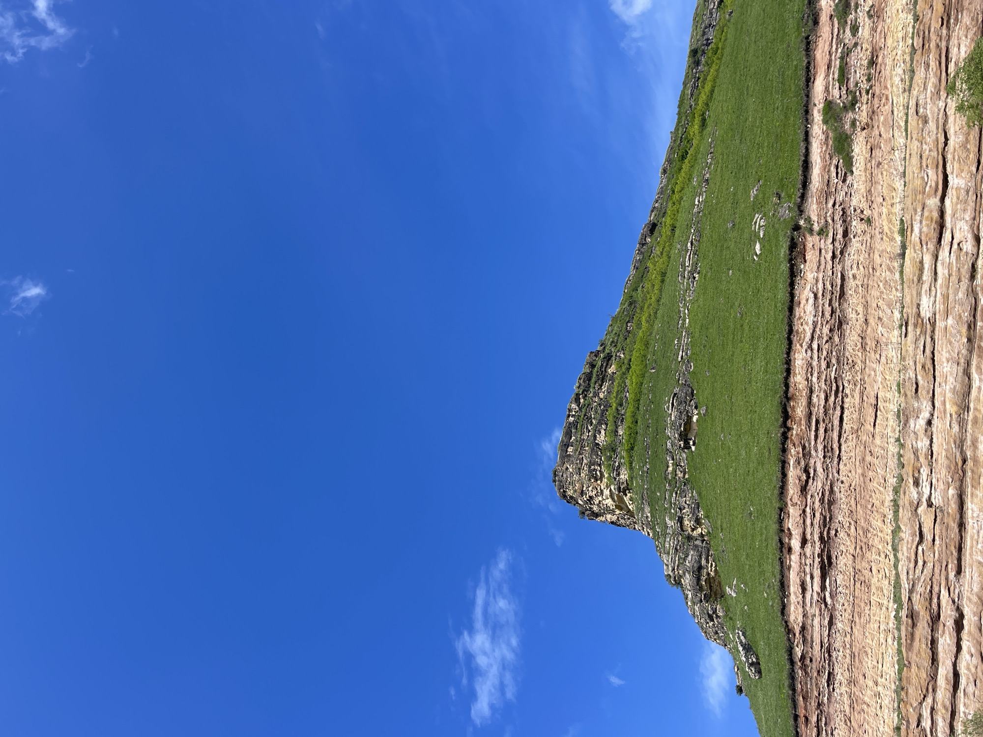 The image shows a landscape with a clear blue sky and a cliff face. The sky is predominantly blue with scattered wisps of white clouds. The cliff face is covered in green vegetation at the top, transitioning to bare rock layers on the lower portion. The rock layers are striated and appear to be composed of sedimentary rock. The image appears to be taken in a rural setting, although specific details about the location in Красный Курган, Russia are not evident. There are no visible people or signs of human activity in the immediate scene.