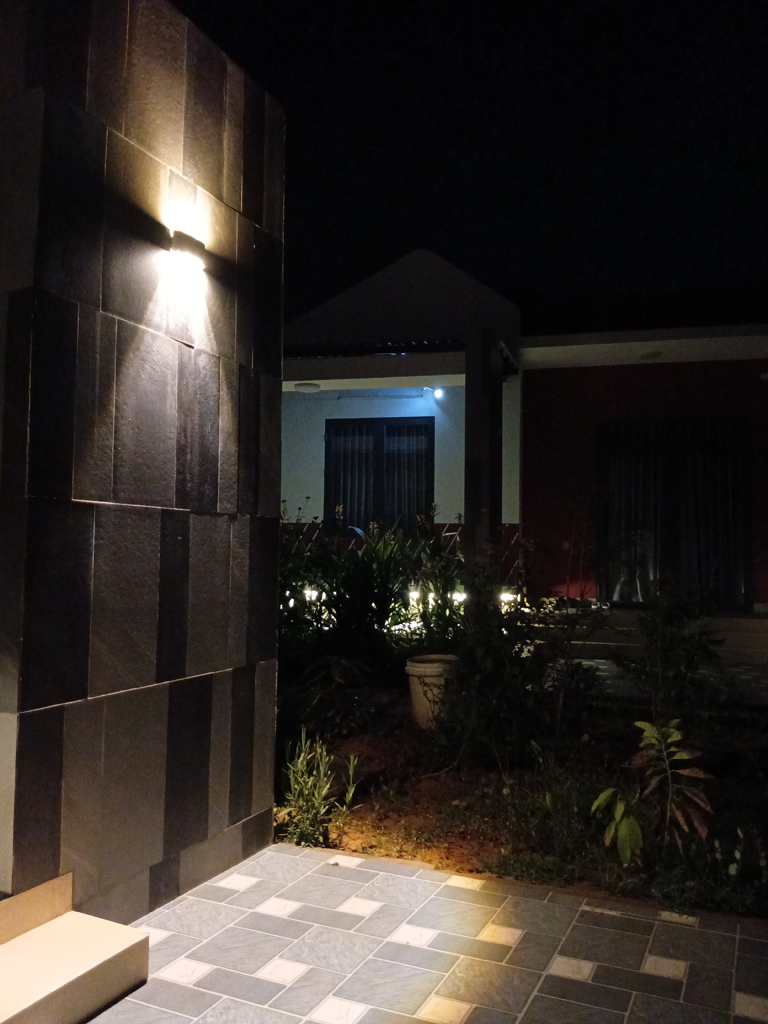 At night in Mũi Né, Vietnam, a modern house with exterior lighting is partially visible. A wall constructed of dark grey stacked stone is illuminated by a wall-mounted light fixture, casting a bright glow. In the background, a building with a white façade and a visible window can be seen, partially obscured by garden foliage. Small ground-level lights illuminate the garden, which features various plants. A paved area in the foreground is constructed with grey and white patterned tiles, reflecting some of the light. The overall scene is dark, emphasizing the artificial lighting and modern architectural elements.