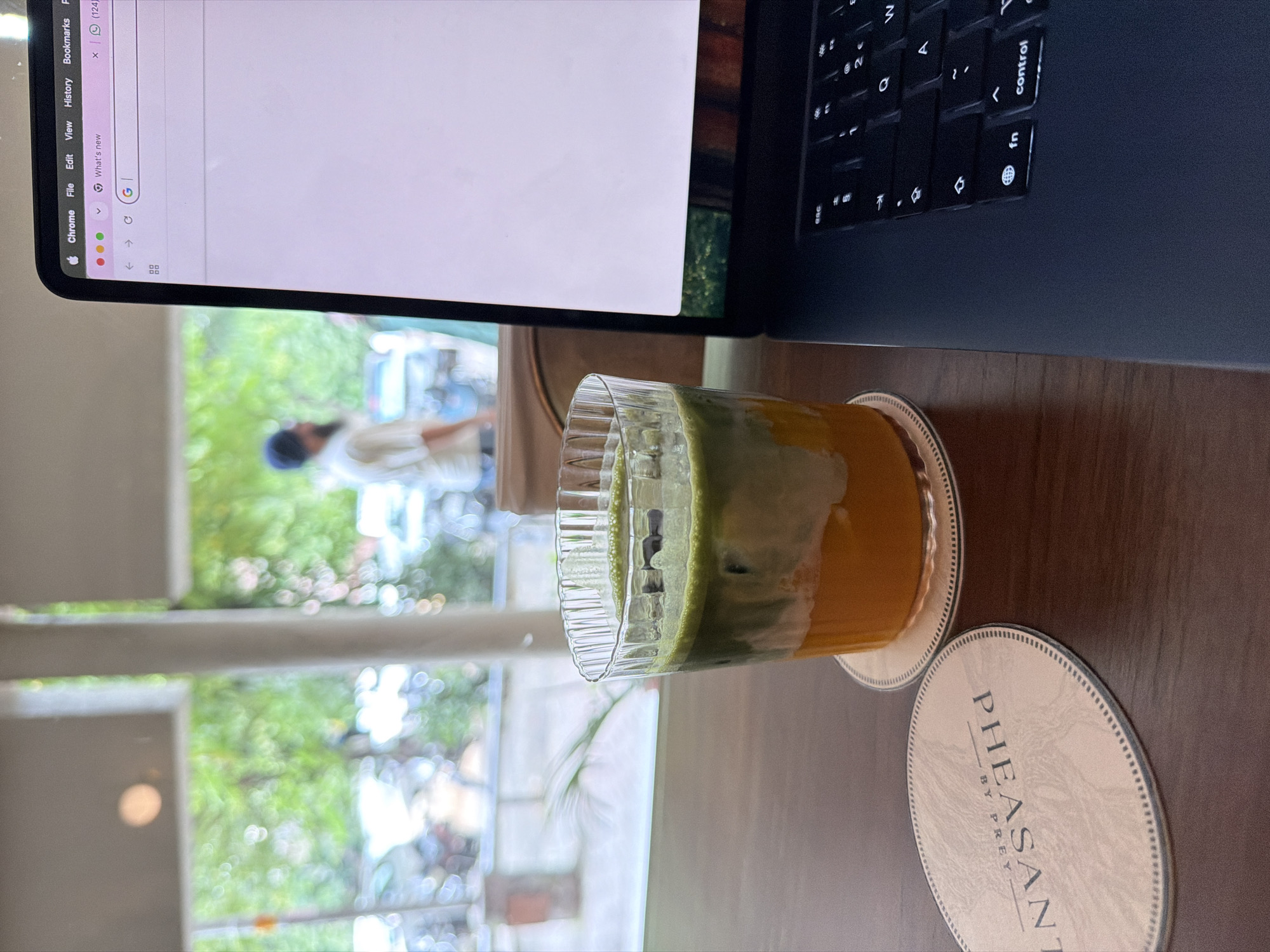 An eye-level, close-up shot shows a layered drink in a fluted glass on a coaster on a dark wooden table. The drink has an orange base and a green layer on top with ice cubes. A laptop is open to the right, displaying a Chrome browser window. A light-colored coaster with the text "PHEASANT BY PREY" is visible to the left of the drink. In the background, a blurred view through a window shows greenery and people. This scene is in Chandigarh, India.