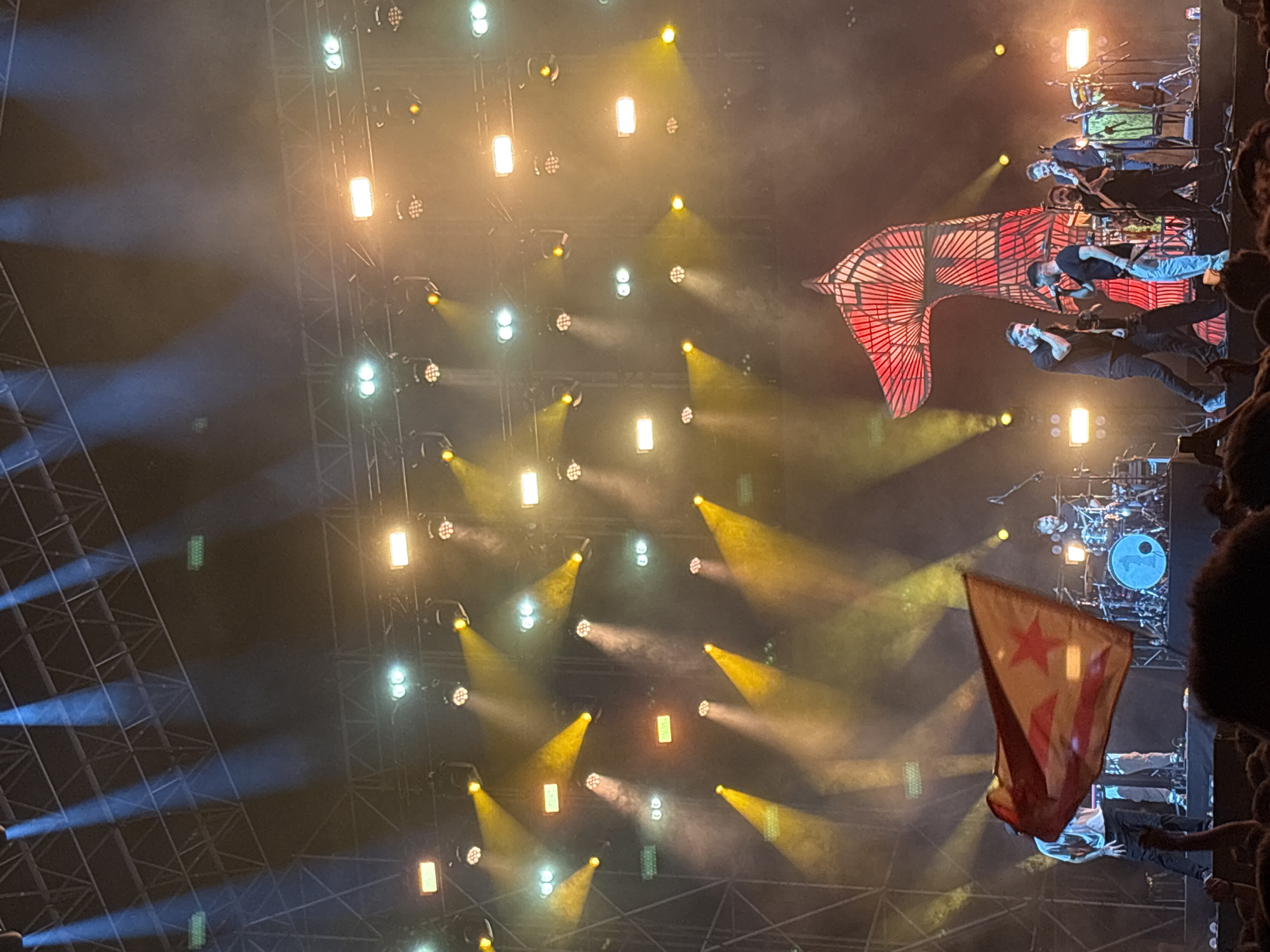 The image depicts a live music concert, potentially in Cunit, Spain. The stage is illuminated with numerous spotlights, some white and some yellow, creating a smoky, theatrical atmosphere. On stage, a band is performing, with at least three visible members: two vocalists at the front and others behind them, including a drummer behind the center. A large red flag is displayed on the stage, along with a smaller flag with a star on it being held in the crowd in front of the stage. Part of a metal stage structure and crowd members are visible on the right side of the image. The focus appears to be on the band and the stage lighting.