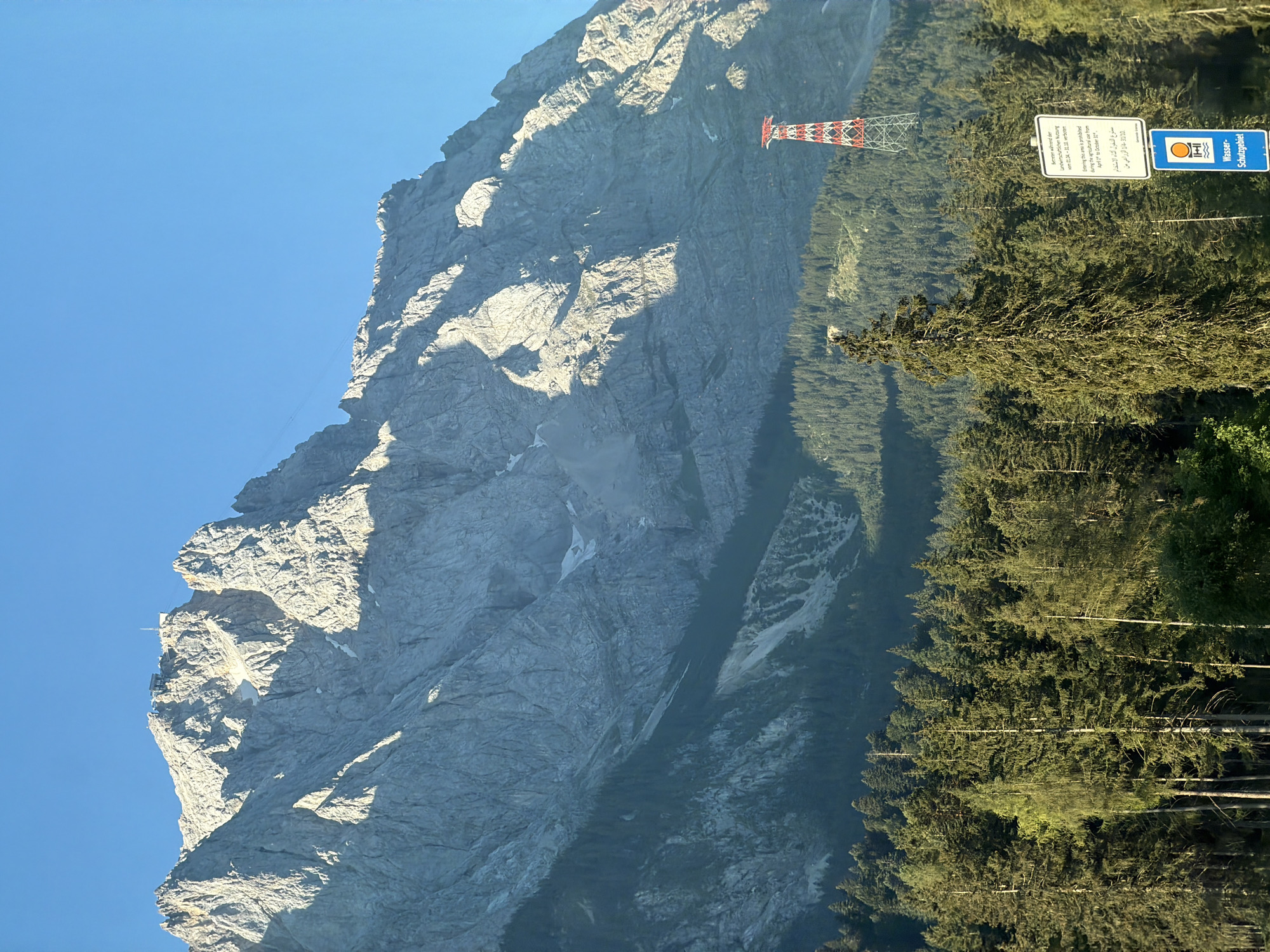 This image depicts a majestic mountain landscape under a clear blue sky. In the foreground, a dense forest of evergreen trees stretches across the frame, with some trees taller than others, reaching towards the sky. The sunlight filters through the trees, casting shadows and highlighting the textures of the needles and bark.
Dominating the background is a massive, rugged mountain range. Its steep slopes are etched with rock formations, crevices, and patches of snow. The sunlight accentuates the contours and dramatic shadows of the mountain, giving it a formidable presence. A small building or structure is visible on the highest peak, suggesting a human presence at the summit, likely a weather station or a visitor facility.
To the right of the mountain, a red and white lattice tower stands tall, its geometric structure contrasting with the natural forms of the mountain and forest. Two signs are visible near the tower; one is a white rectangular sign with text in multiple languages, indicating regulations or information, and below it, a blue sign with a white symbol and the text "Wasser-Schutzgebiet" (Water Protection Area).
The overall scene evokes a sense of natural grandeur and the majesty of the alpine environment. The location is identified as Grainau, Germany, which is known for its proximity to the Zugspitze, Germany's highest mountain, and its surrounding natural beauty.