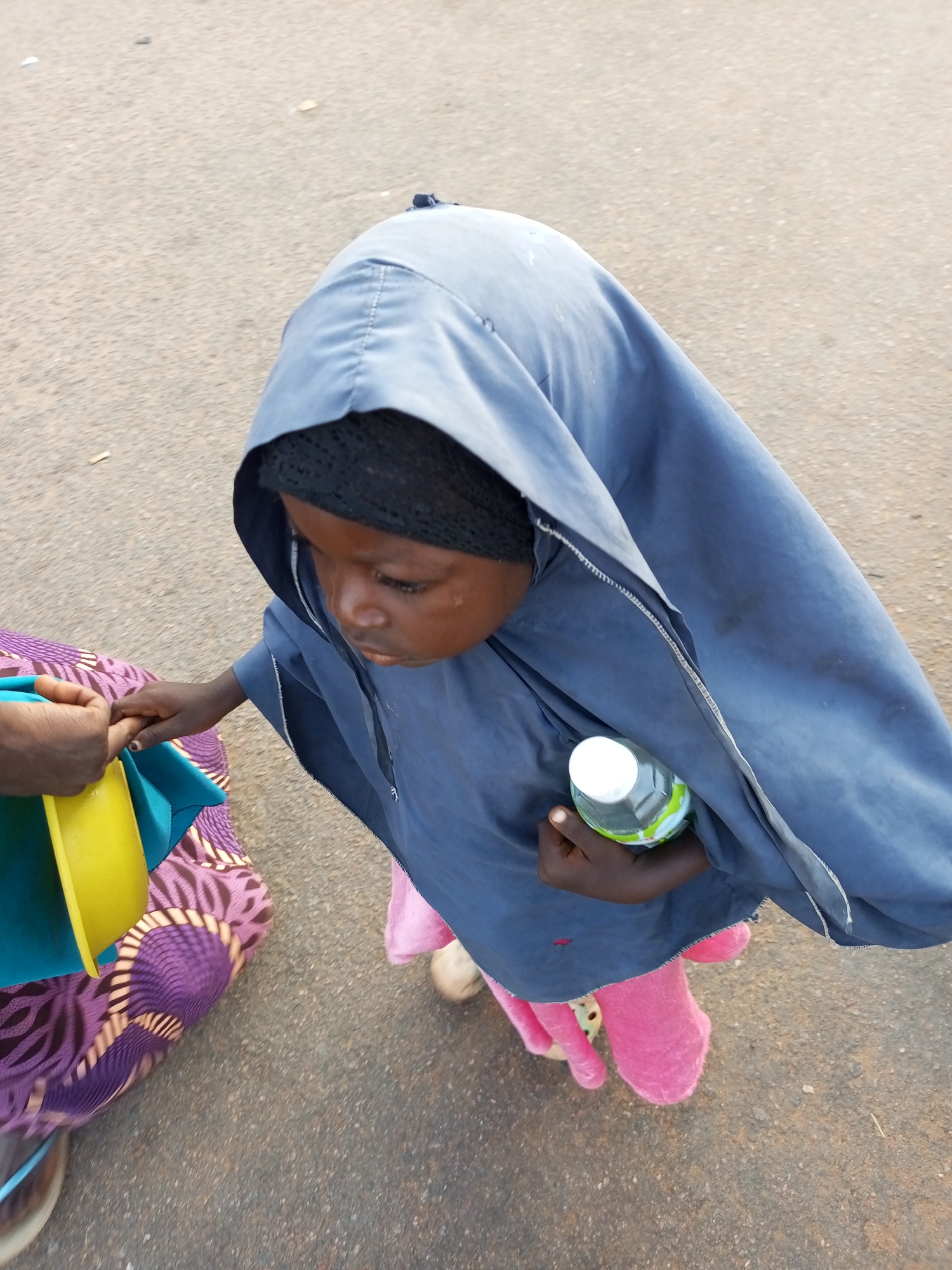The image shows a child wearing a blue head covering and pink pants, holding a bottle of water and a yellow bowl, standing on a paved surface that appears to be a street or sidewalk in Gombe, Nigeria. The child's face is visible, looking downwards. Another person's hands are visible on the left side of the image, wearing a purple and pink patterned garment and holding onto the child's hand and the yellow bowl. The environment is an outdoor urban setting with asphalt ground.