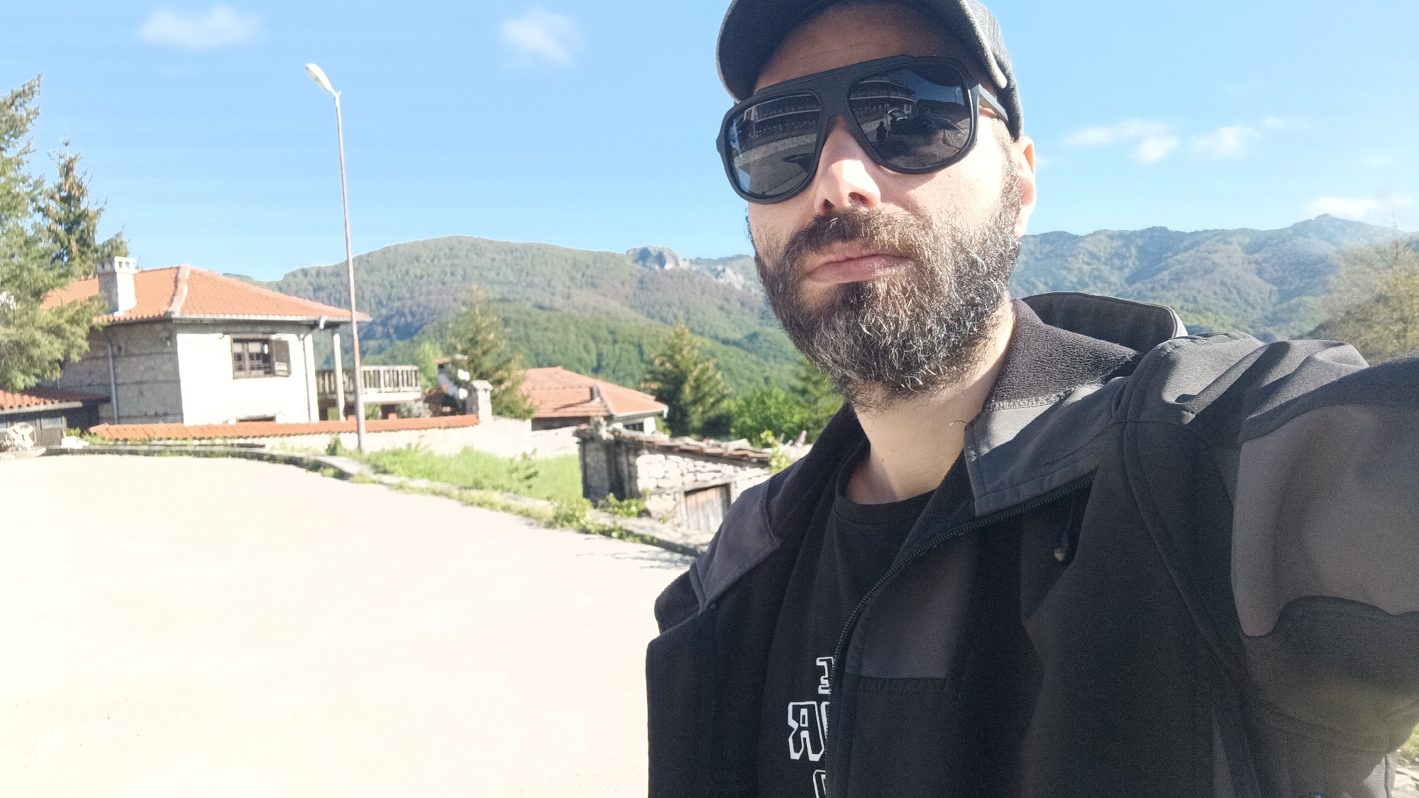 The image is a selfie of a man with a beard and sunglasses, wearing a cap and black jacket. In the background are houses with red roofs, a lamppost, and forested mountains under a partly cloudy sky. The location is inferred to be in the city of Vrata, Bulgaria, based on the prompt. The man appears to be standing on a paved surface, likely a street or road. The photo was probably taken during the day, based on the lighting.