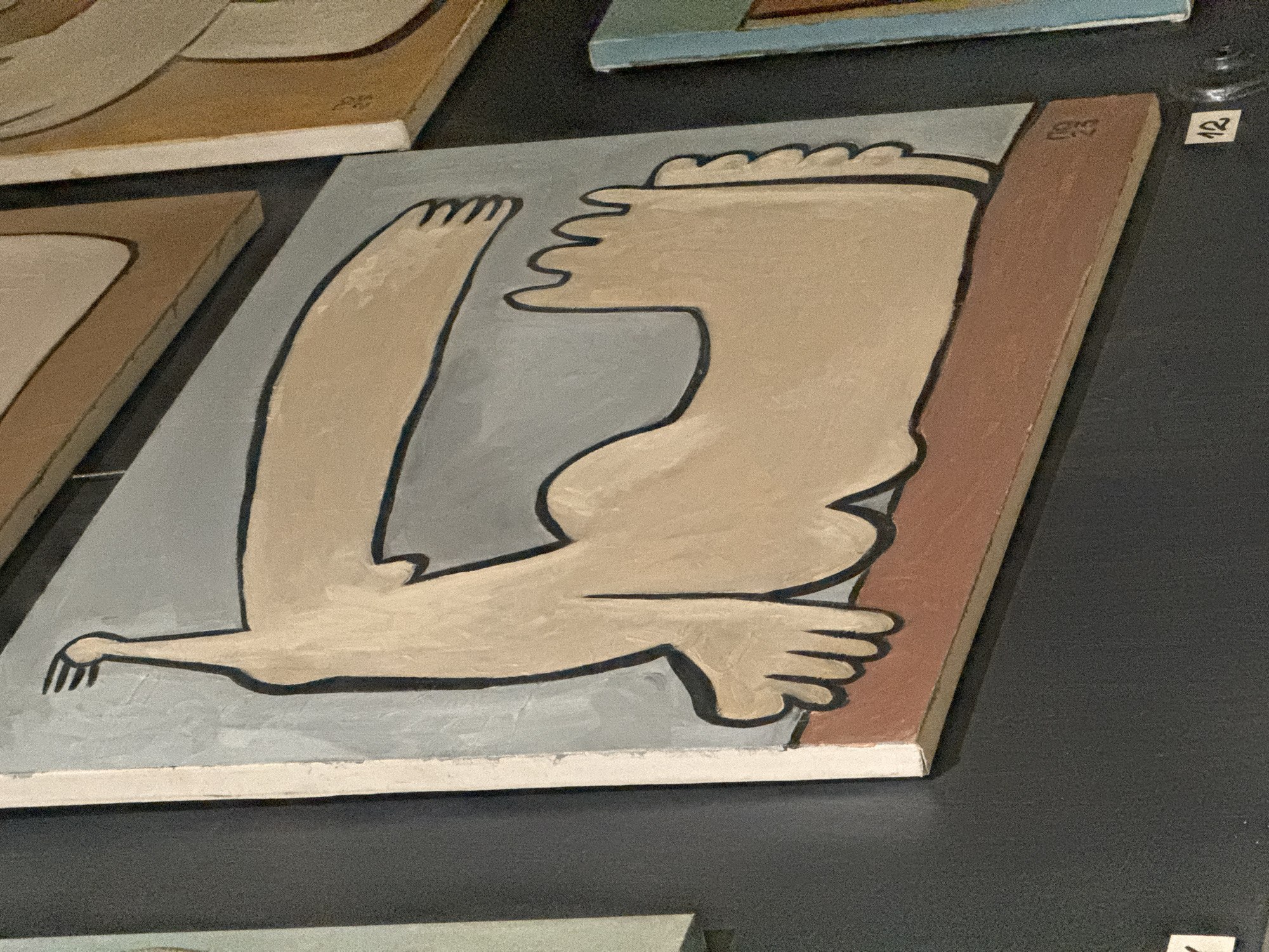 This media file presents a close-up view of a modern, abstract painting displayed in what appears to be an art exhibition or gallery in Varna, Bulgaria. The central artwork is a rectangular panel or canvas depicting stylized human limbs.
The painting features a light beige or tan color for the limbs, outlined with thick black lines, against a light grey-blue background. A long, slender arm with a distinct hand and exaggerated fingers extends across the lower left part of the canvas. Above and to the right, a larger, more robust form suggests a torso connected to a leg, with clearly defined toes at the very top edge of the panel. A reddish-brown vertical strip frames the right side of the painted composition. The art style is graphic and simplified, reminiscent of early 20th-century avant-garde movements.
The painting is mounted against a dark, possibly black, wall. Other artworks of a similar style are partially visible in the blurred foreground and background, indicating a larger display. On the dark wall, to the right of the main painting, a small white label with the number "12" printed in black is visible. Faintly visible on the reddish-brown strip within the painting itself is a small, faded marking, possibly "25" or "ES." The setting is indoors, under artificial lighting, focusing on the visual characteristics of the artwork.