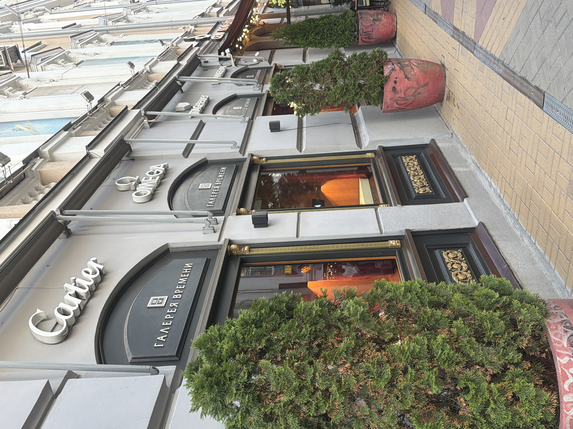 The image, oriented vertically, captures a section of a city street facade, focusing on several high-end storefronts. The building features light-colored, possibly grey, architectural details on its upper levels, including ornate cornices and corbels.
At street level, the scene is dominated by well-maintained evergreen shrubs housed in large, weathered red terracotta planters. These planters sit on a sidewalk paved with interlocking beige and light brown tiles, alongside a metal drainage grate.
The storefronts themselves exhibit a luxurious aesthetic, with dark-colored frames and gold-toned decorative elements around the display windows and entryways. Prominent signage identifies the businesses. The brand "Cartier" is visible in elegant silver letters, and further along the facade, the brand "OMEGA" is also displayed in silver. Adjacent to both brand names, and positioned above the entrances, are signs in Cyrillic script reading "ГАЛЕРЕЯ ВРЕМЕНИ," which translates to "Gallery of Time." Each "ГАЛЕРЕЯ ВРЕМЕНИ" sign is accompanied by a small emblem featuring what appears to be Roman numerals (possibly VI or XII) within a square.
The shop windows reflect interior lighting, indicating well-illuminated display areas. The overall impression is that of a high-end shopping district. The scene appears to be set during the daytime, with soft, ambient light suggesting either an overcast sky or indirect sunlight. No people are visible, and no specific activity is taking place, presenting a static view of the urban environment. The presence of Cyrillic text and the provided context indicate the location as Rostov-on-Don, Russia.