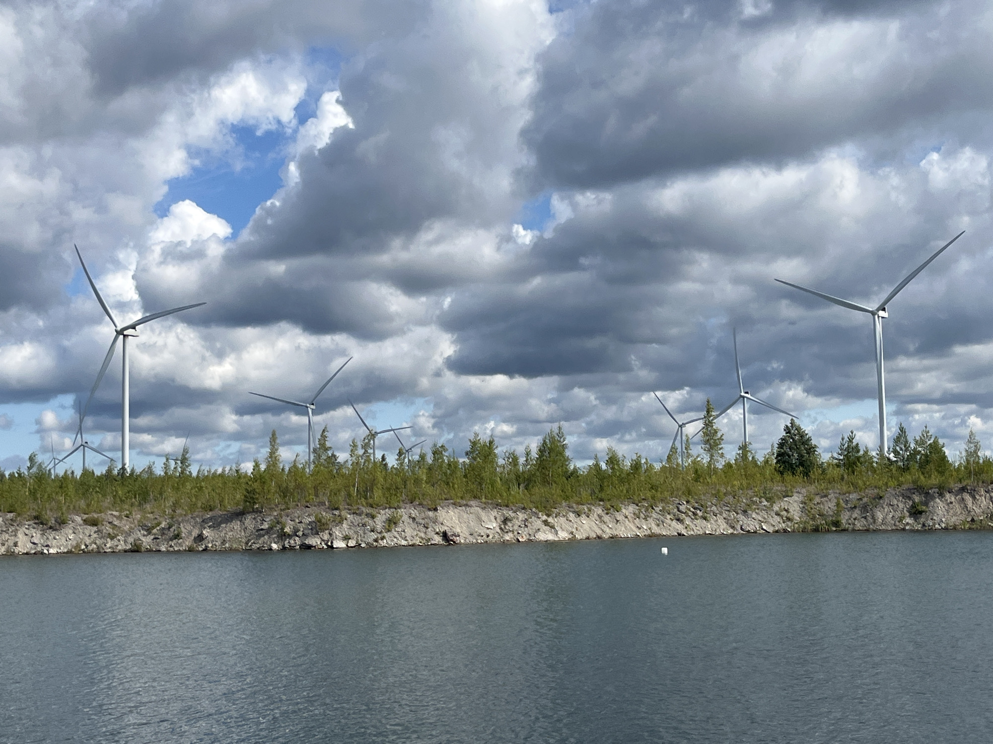The image depicts a body of water in the foreground, with a rocky, vegetated shoreline beyond. Several wind turbines are visible on the distant horizon, silhouetted against a cloudy sky. The wind turbines are tall, white structures with three blades each, and they appear to be in operation. The shoreline is lined with green trees and shrubs, and there are some scattered rocks and debris on the ground. The sky is filled with dramatic, grey clouds, with patches of blue sky visible between them. The overall scene suggests a rural or industrial landscape, likely used for renewable energy generation. The location is stated to be Ojamaa, Estonia, which is known for its wind farms.