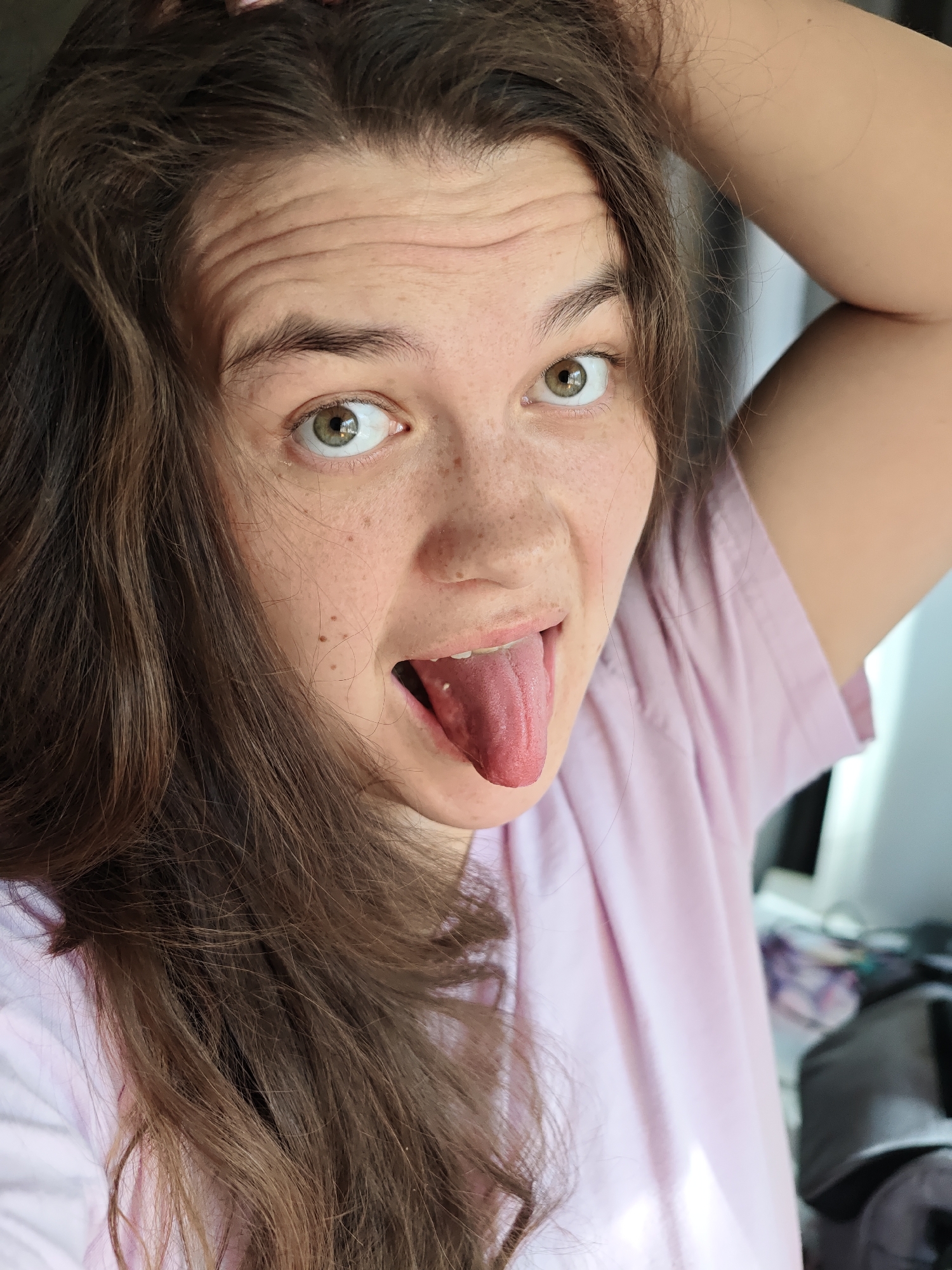 A woman with light skin, brown hair, and green eyes is sticking her tongue out at the camera. She has freckles on her face. She is wearing a light pink shirt. Her hair is long and wavy. Her right arm is raised. The background is blurred, but it appears to be an indoor setting with a window and some clothing or other objects. There is no indication of the location being Chaikovsky, Russia, within the image.