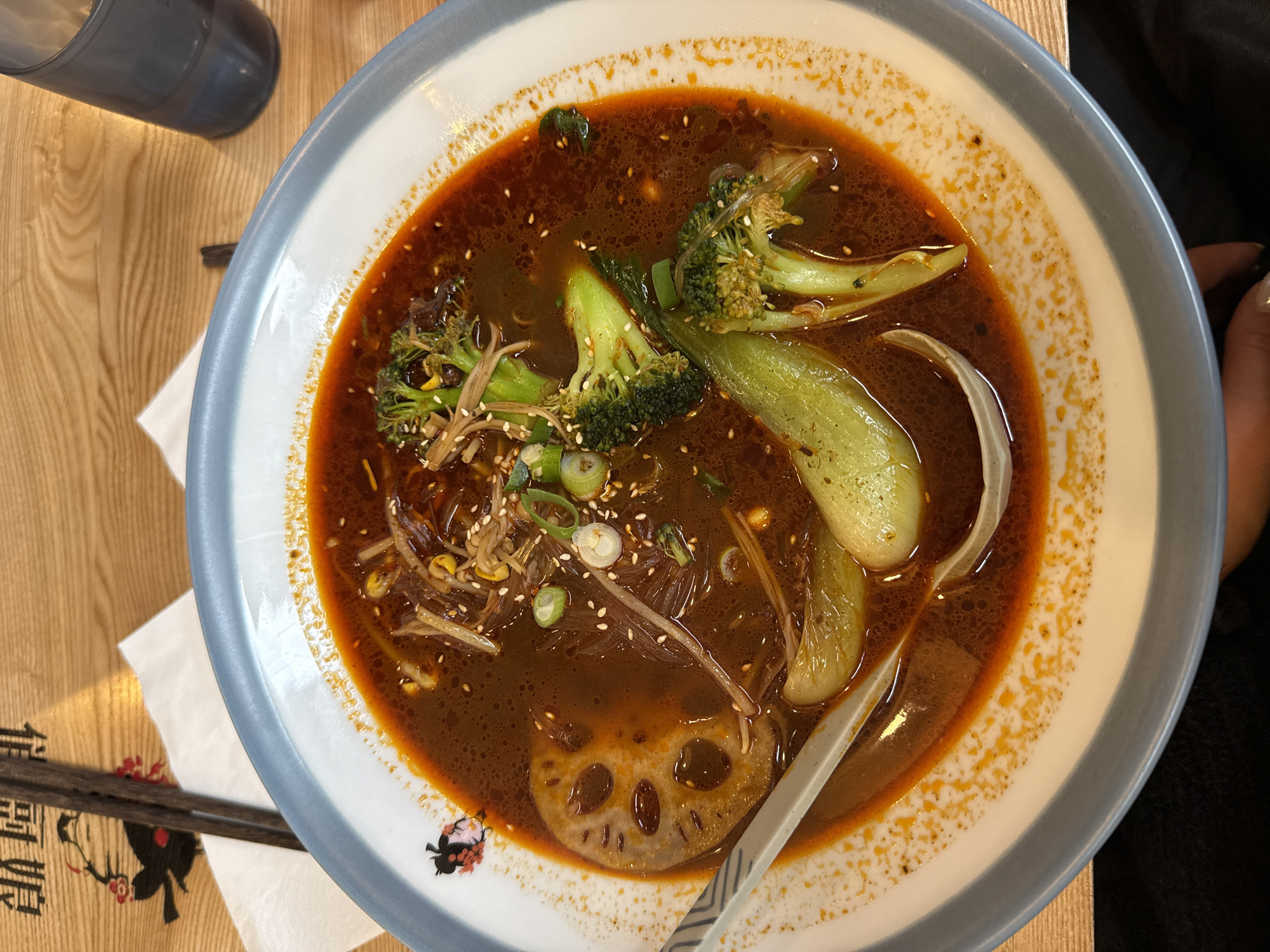 A top-down, eye-level shot presents a bowl of Asian cuisine. The bowl, with a white interior and a light blue rim, contains a dark brown broth filled with various ingredients. Broccoli florets, bok choy, thin noodles, sesame seeds, and sliced scallions are visible. A lotus root slice rests on the bottom left. A plastic spoon is present in the bowl.
To the left of the bowl are chopsticks on top of a napkin with graphics and text in a foreign language. Further left is a partially visible glass or water bottle. The table beneath the bowl has a light wooden surface. A portion of a person's arm is visible on the right side of the image. There is no explicit information about the location in Paris, France.