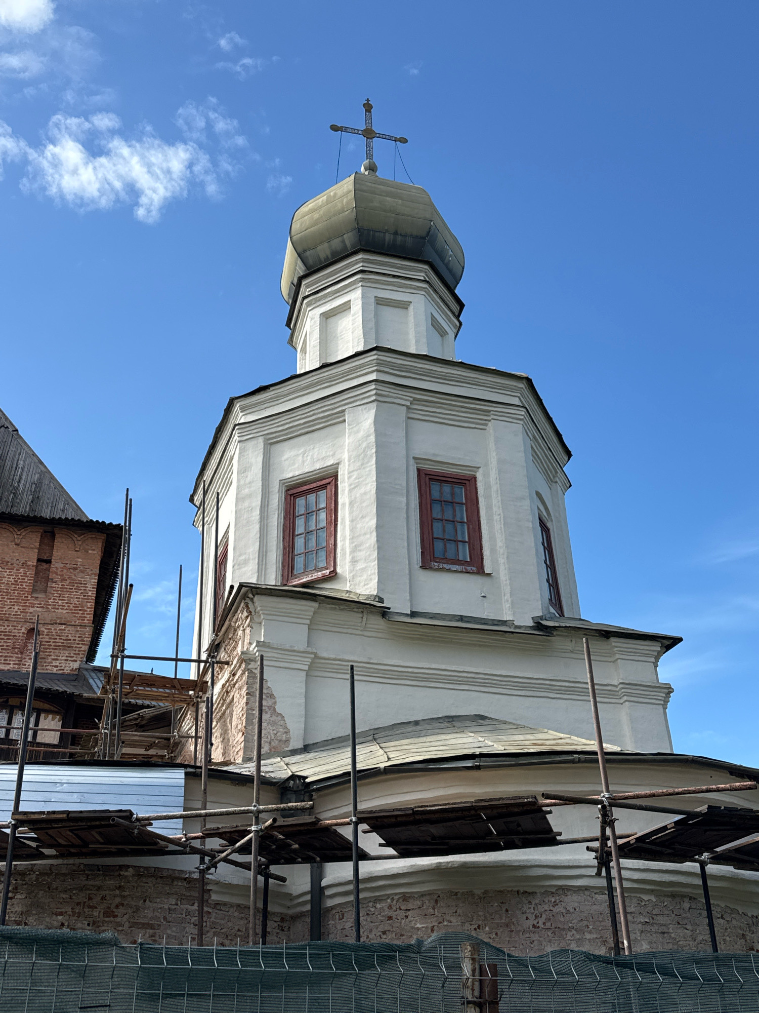 The image shows a close-up, upward-angle view of a white, octagonal church bell tower topped with a green, onion-shaped dome and a cross. The bell tower has red-trimmed windows and ornate cornices. Scaffolding surrounds the lower portion of the bell tower and the adjacent brick building, indicating ongoing construction or renovation. The sky is clear blue with a few wispy clouds. This scene is located in Veliky Novgorod, Russia, specifically at the Yaroslav's Court.