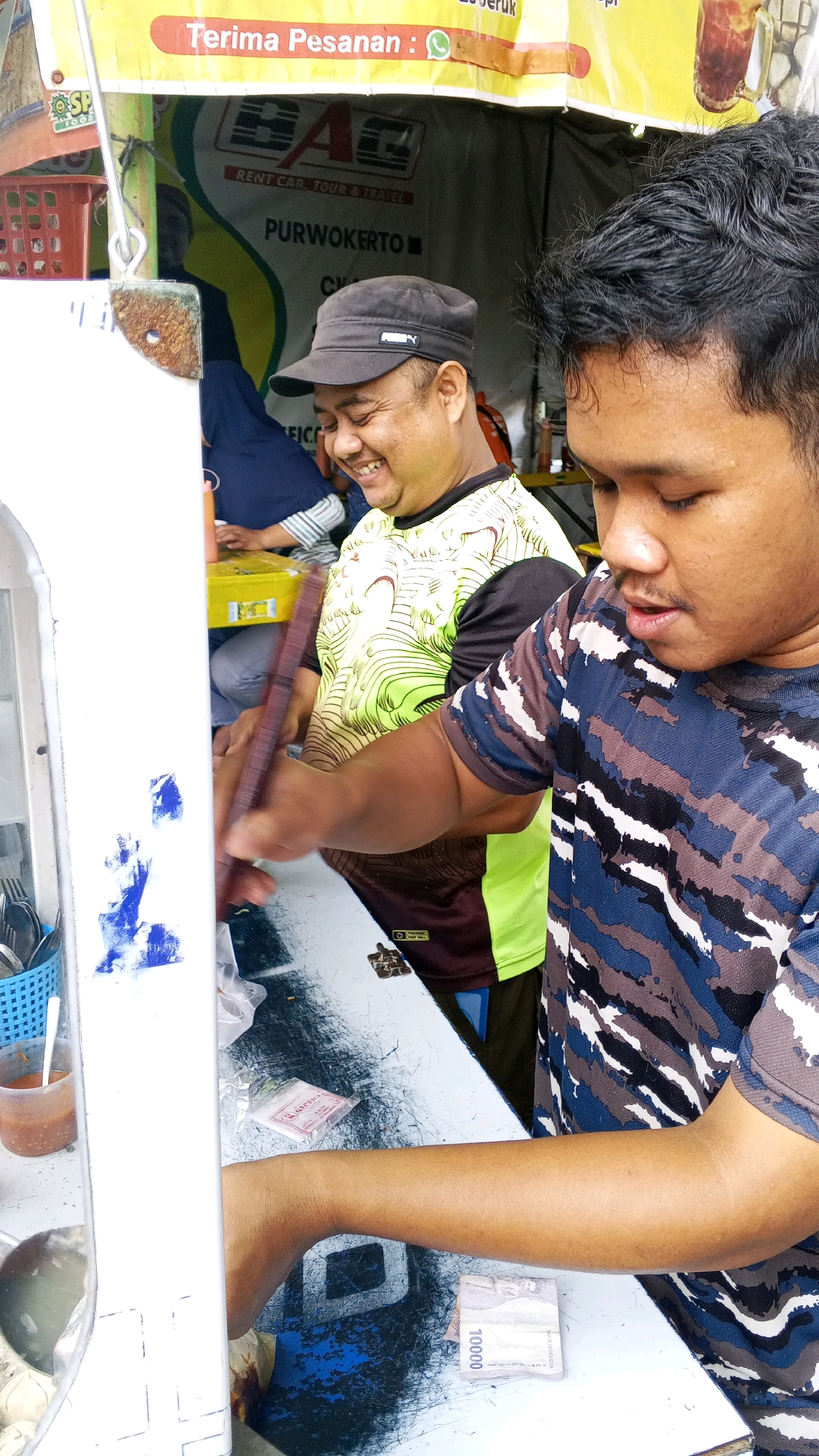 Two Indonesian men are at a food stall. The man on the left, wearing a black cap and a lime green and black shirt, is smiling and appears to be interacting with someone off-camera. The man on the right, wearing a camouflage shirt, is focused on a task in front of him. The stall has a yellow banner at the top with Indonesian text that reads "Terima Pesanan" (Take Orders) and a symbol of a smartphone, indicating an order can be placed via phone. Below that, a banner displays "BAG RENT CAR, TOUR & TRAVEL PURWOKERTO". The table in front of the men has various items including packets, utensils, and Indonesian currency. The background shows the interior of the stall with more signage and baskets. The scene depicts a typical street food vendor setting in Semarang, Indonesia.