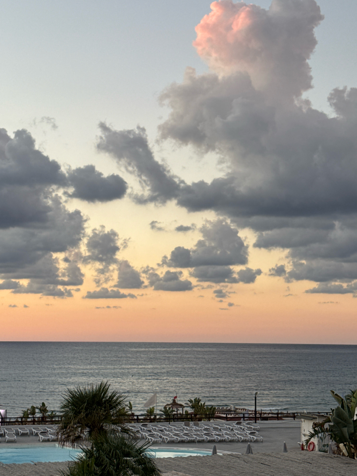 The image depicts a beach scene in Capaci, Italy, at dusk. The sky is filled with dramatic clouds, some tinged with pink and orange from the setting sun, while others are dark gray. The sea is visible in the middle ground, with calm waves. The foreground shows a beach with rows of empty lounge chairs, suggesting the end of the day. Palm trees and tropical plants are scattered across the sand. A swimming pool is also visible. The overall atmosphere is serene and tranquil.