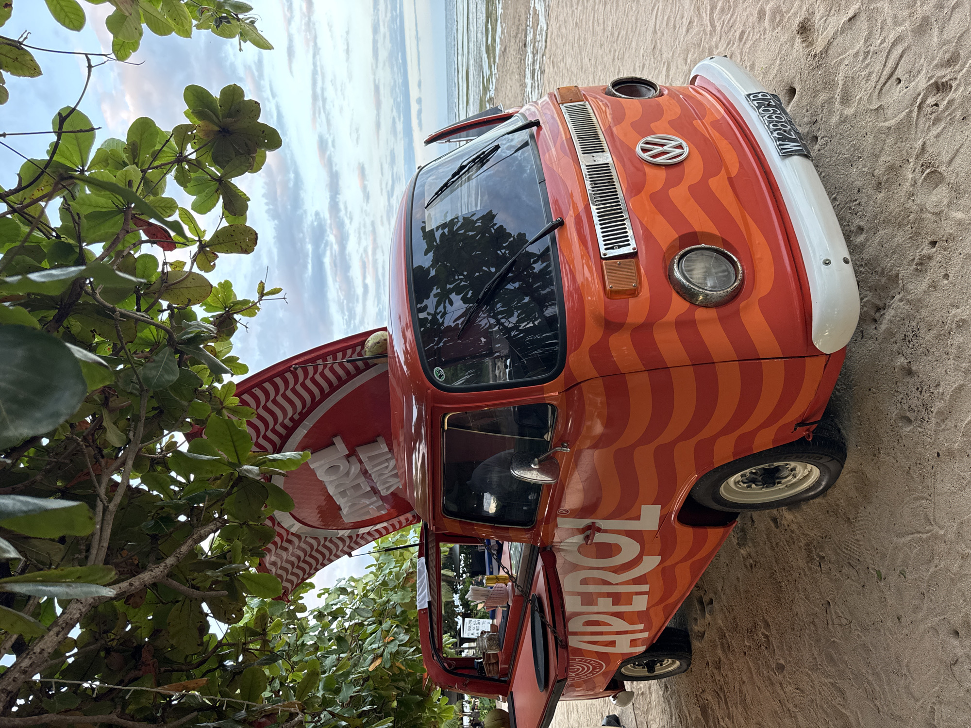 The image shows an orange and white Volkswagen van parked on a sandy beach, likely in Kuta Selatan, Indonesia. The van features an Aperol advertisement along the side, suggesting it might be used as a mobile bar or vendor. The van's license plate reads "W 2866 GB". Part of the vehicle is shaded by a tree with green leaves. In the background, the sea meets a cloudy sky. The sand is marked with footprints, indicating human activity. The angle of the image is slightly tilted.