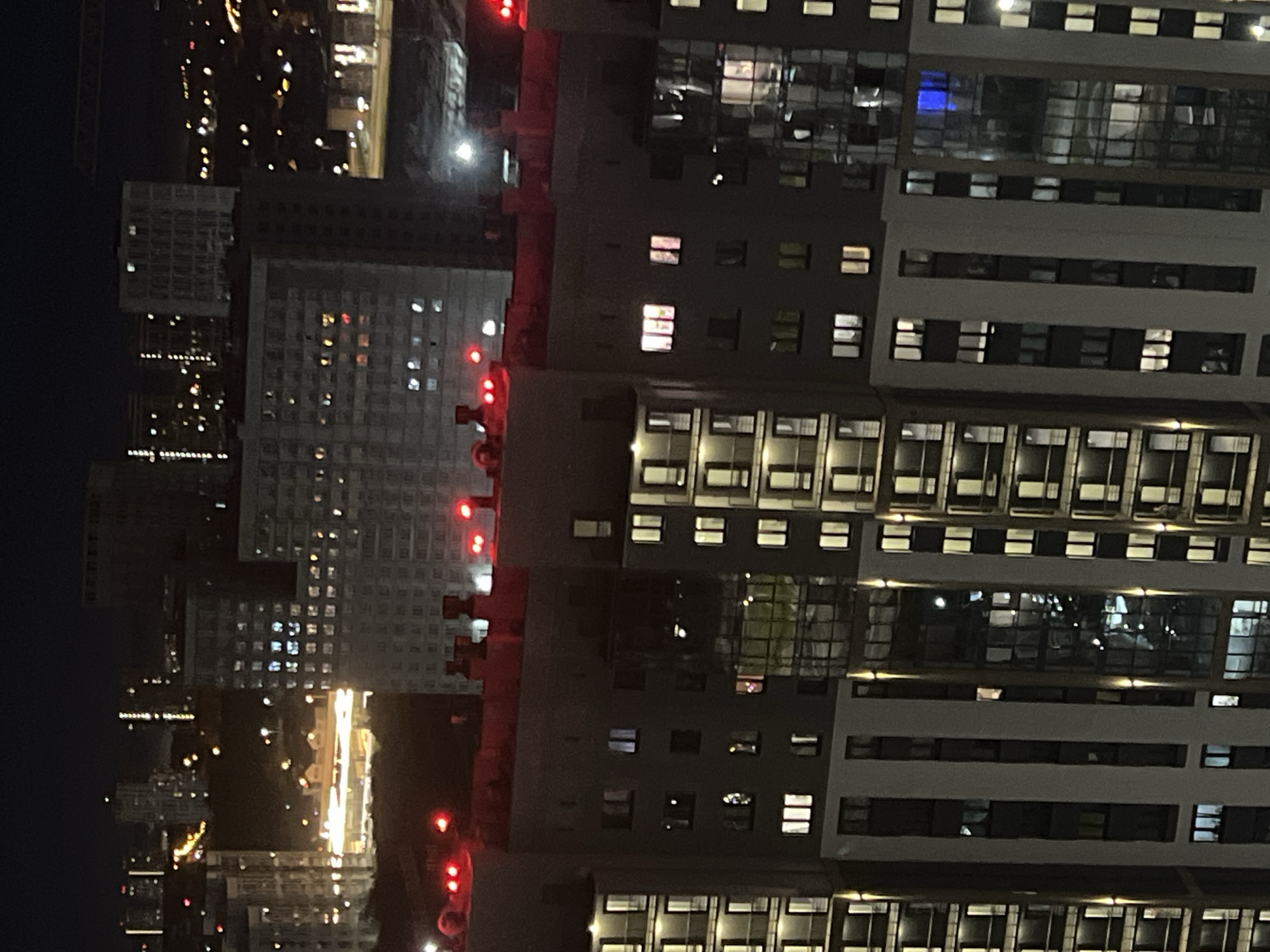 The image shows a night-time aerial view of a residential area in Yekaterinburg, Russia, likely in the vicinity of the "Ural Towers" or similar modern high-rise apartment complexes. The foreground is dominated by the illuminated windows and dark facades of several tall apartment buildings, showcasing a grid-like pattern of lights against the night sky. Some buildings have balconies visible, and red warning lights are present on the rooftops of some structures. In the mid-ground, more apartment buildings and some construction cranes are visible, suggesting ongoing development or a recently developed area. The background reveals a city skyline with distant lights, indicating the presence of other urban structures and a wider cityscape. The overall scene depicts a densely populated urban environment at night, characterized by a high concentration of residential buildings. There is no clear indication of any specific events or activities beyond the general depiction of urban life.