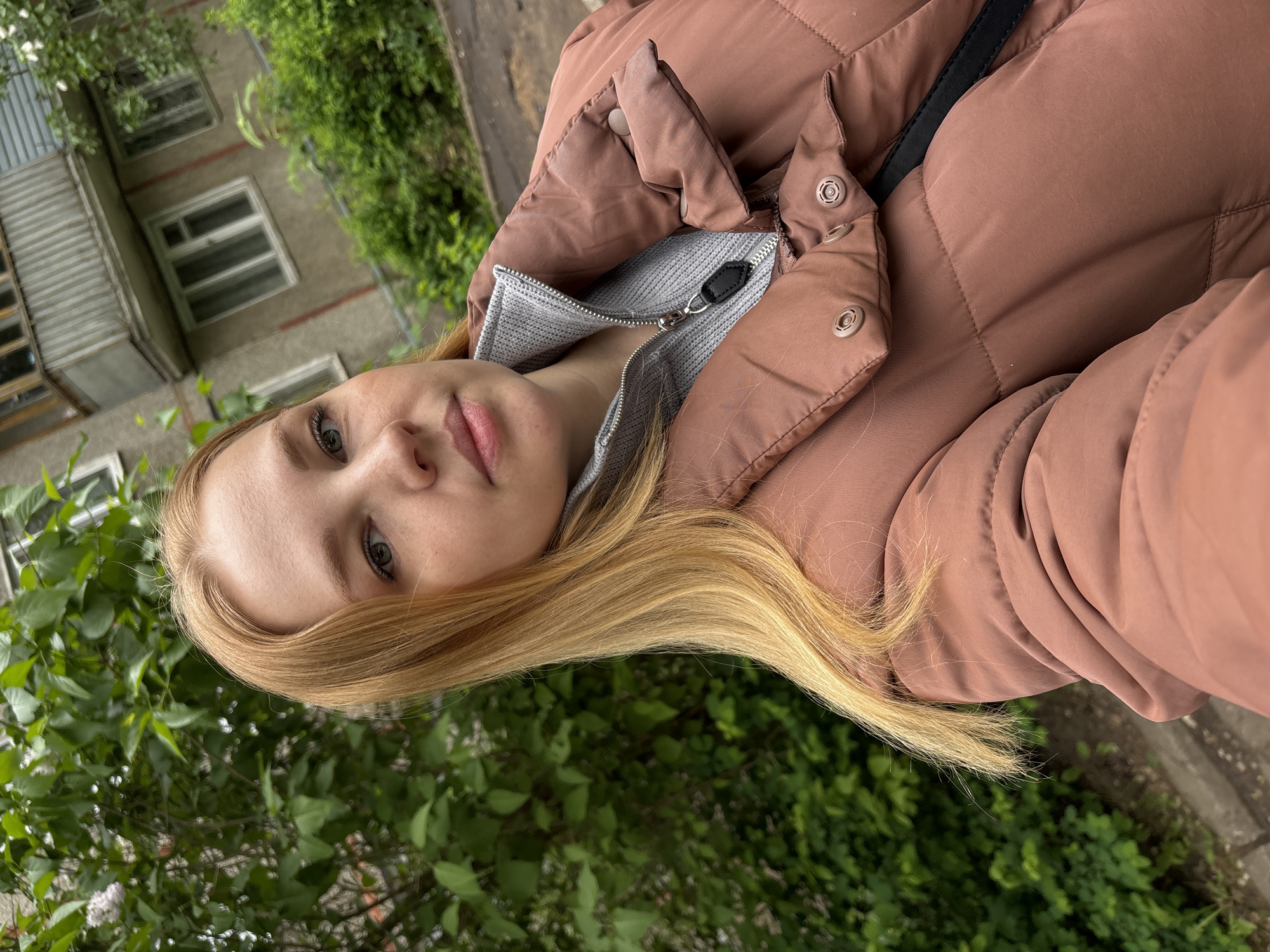 A young, light-skinned woman with blonde hair and light blue-green eyes is pictured in an outdoor setting. She is wearing a brown puffer jacket over a light grey top with a partial zipper. The jacket is partially open, revealing the grey top underneath. A black strap, possibly for a bag, is visible on her shoulder. The background includes green foliage, possibly trees or bushes. A multi-story building with white-framed windows is visible in the upper-left corner of the frame. The image is taken at an angle, with the camera pointing slightly upwards towards the subject. The location is identified as Rusanovo, Russia.