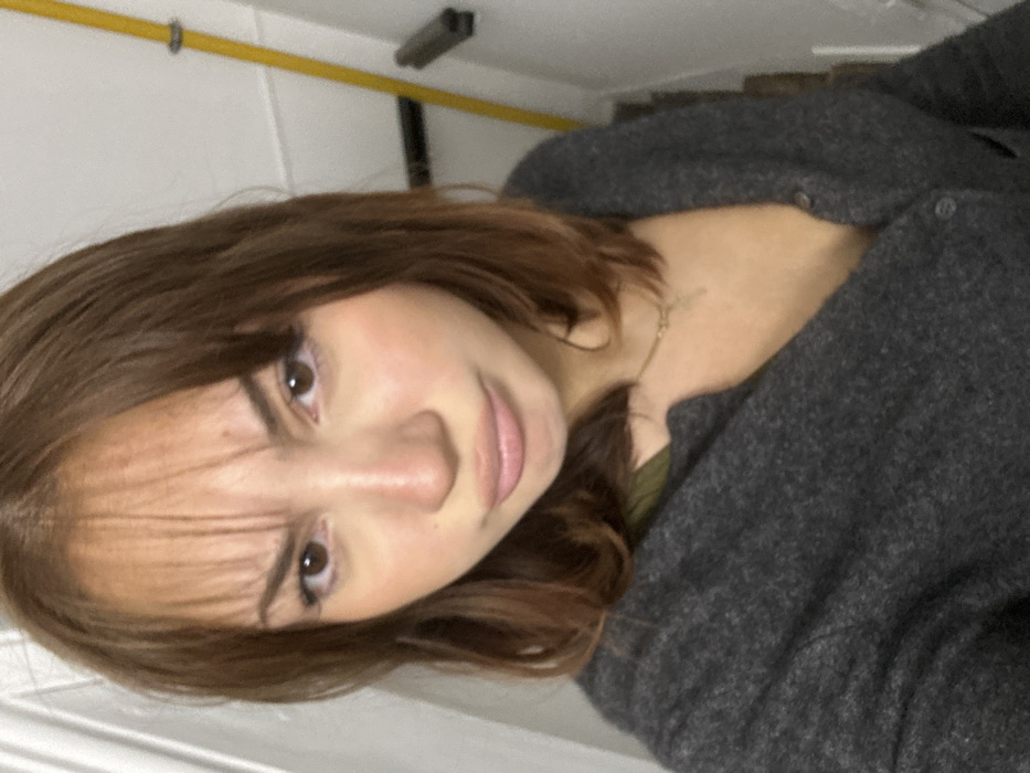 A young adult female is centered in an indoor, chest-up portrait. She has medium-brown hair with bangs, fair skin, and brown eyes, with her lips slightly pouted and eyebrows subtly furrowed. She wears a dark grey knit cardigan, partially unbuttoned to reveal a dark olive-green garment underneath and a thin gold-colored necklace with a small pendant. The background consists of a plain, light-colored wall. A vertical yellow pipe or pole is visible on the right side of the frame, with a dark horizontal bar positioned below it. The precise location within Strasbourg, France, cannot be determined from the visual information provided in the image.