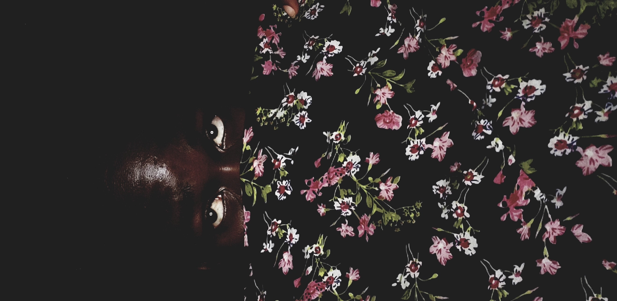 The image shows a close-up, rotated view of a person's face partially obscured by darkness on the left side. The visible skin is dark brown. Two eyes are visible, looking upwards. The right side of the image is dominated by a floral pattern on a black background. The flowers are primarily pink and white, with some green foliage visible. The image doesn't contain enough information to confirm or deny that the photograph was taken in Narayi, Nigeria.