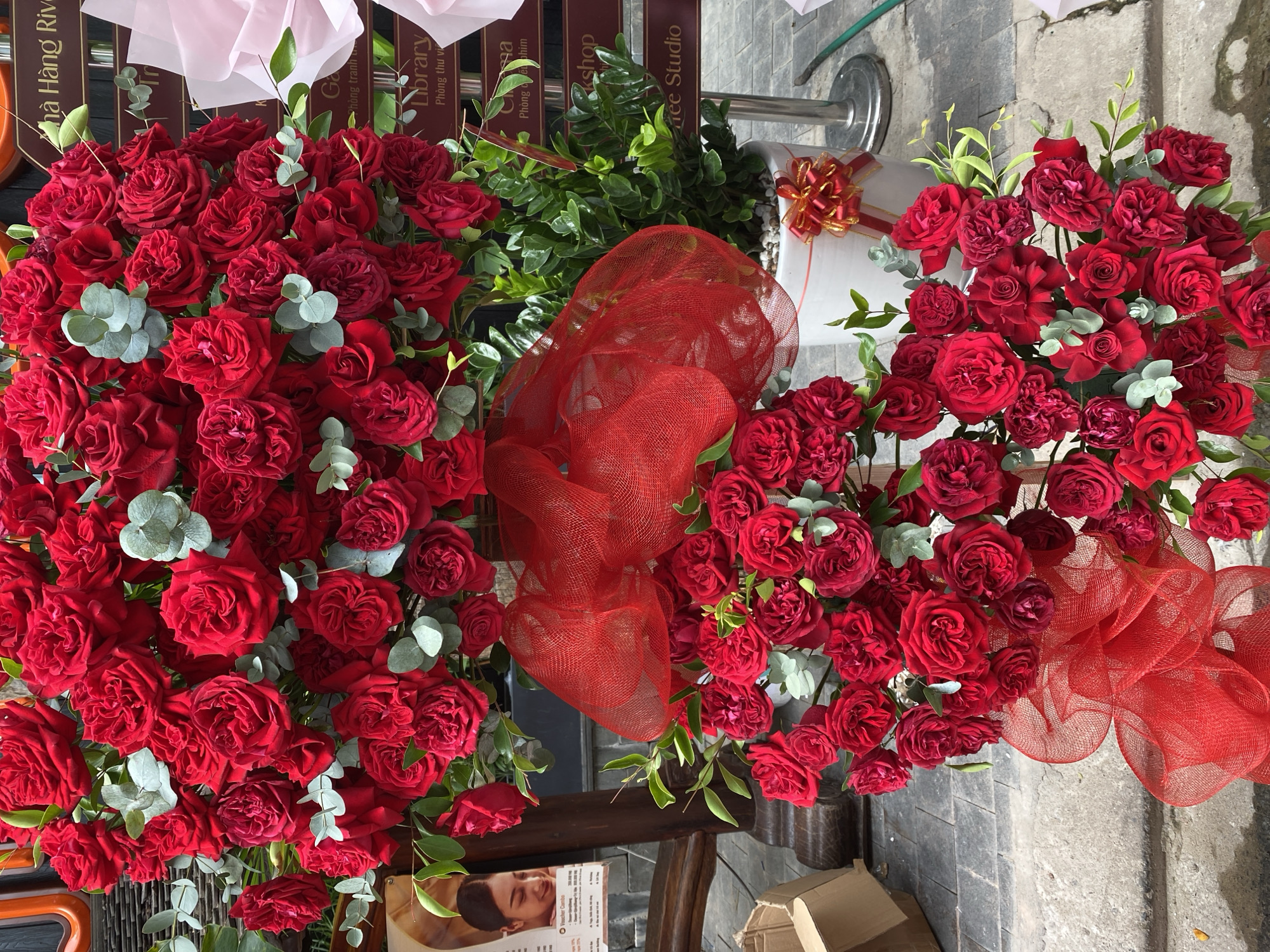 The image depicts a large bouquet of red roses, intricately arranged with eucalyptus leaves and adorned with red netting and bows. The bouquet is positioned outdoors, in what appears to be a street setting in the city of Đà Nẵng, Vietnam. In the background, signs in Vietnamese are visible, including one that reads "Nhà Hàng River". A white potted plant with green foliage sits next to the bouquet. A cardboard box is visible on the left side of the image, and the ground is paved with stone. The lighting suggests it is daytime.