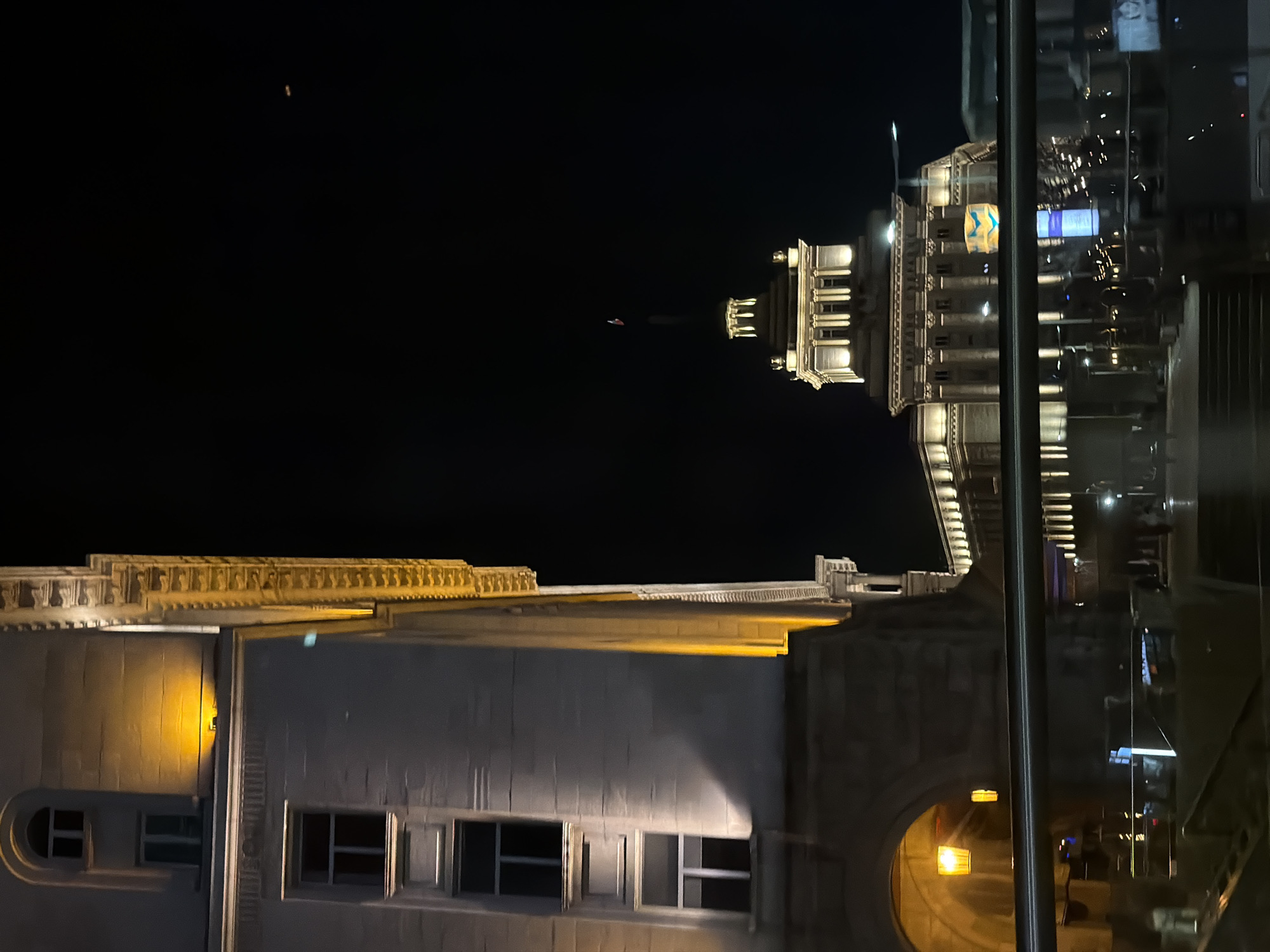 The image shows a nighttime view of buildings in Sofia, Bulgaria. The architecture includes a building with a facade of windows and ornate detailing. The photo is possibly taken through a window, with reflections visible. The buildings are lit up, contrasting with the dark sky. In the background is the building of Largo di Sofia, recognizable by the Ukrainian flag.