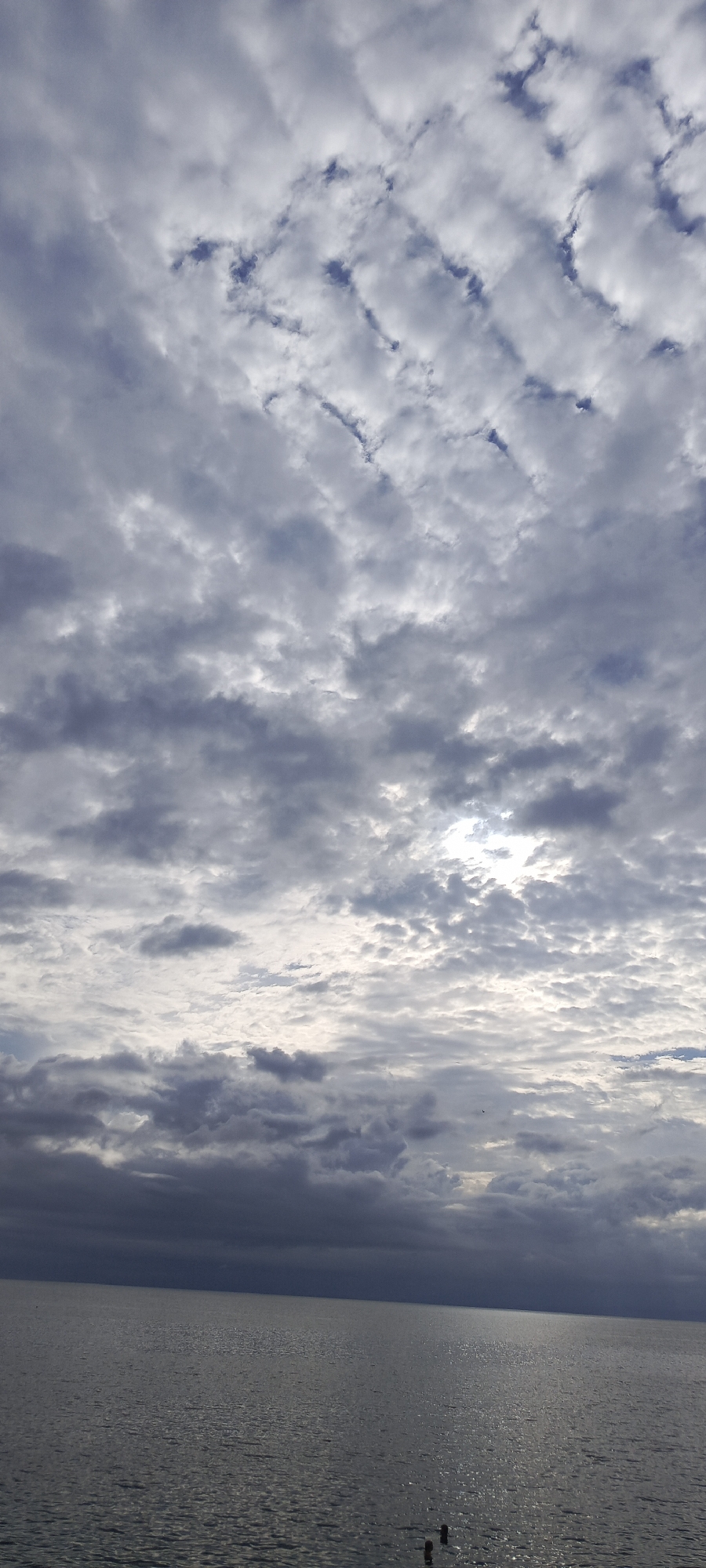 A vast, overcast sky dominates the upper portion of the image, filled with undulating, textured clouds in shades of gray and white. The clouds form a somewhat chaotic yet patterned arrangement, with breaks revealing glimpses of pale blue sky and the sun, which is partially obscured. In the lower half of the image, a calm body of water stretches to the horizon, its surface reflecting the muted light of the sky with a shimmering, textured quality. The water appears dark and still, suggesting a large lake or sea. The horizon line is sharp and distinct, separating the sky from the water. The overall atmosphere is somber and tranquil. No people or specific man-made objects are clearly identifiable within the scene, except for what appear to be two dark, cylindrical posts just above the water's surface near the bottom right corner, possibly indicating a dock or pier. The location is not specified in the image.
