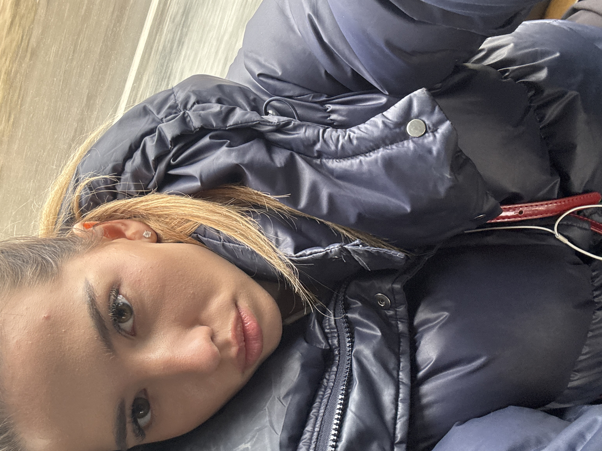A close-up, angled shot shows a woman lying down, largely obscured by a navy blue puffy jacket. Her face, turned slightly to the side, displays light skin, brown hair, and discernible facial features. Part of a red object, likely a belt, is visible near the edge of the jacket. The background appears to be an interior scene with neutral-toned walls. The setting of this image within the city of Кварса, Russia is not discernible.