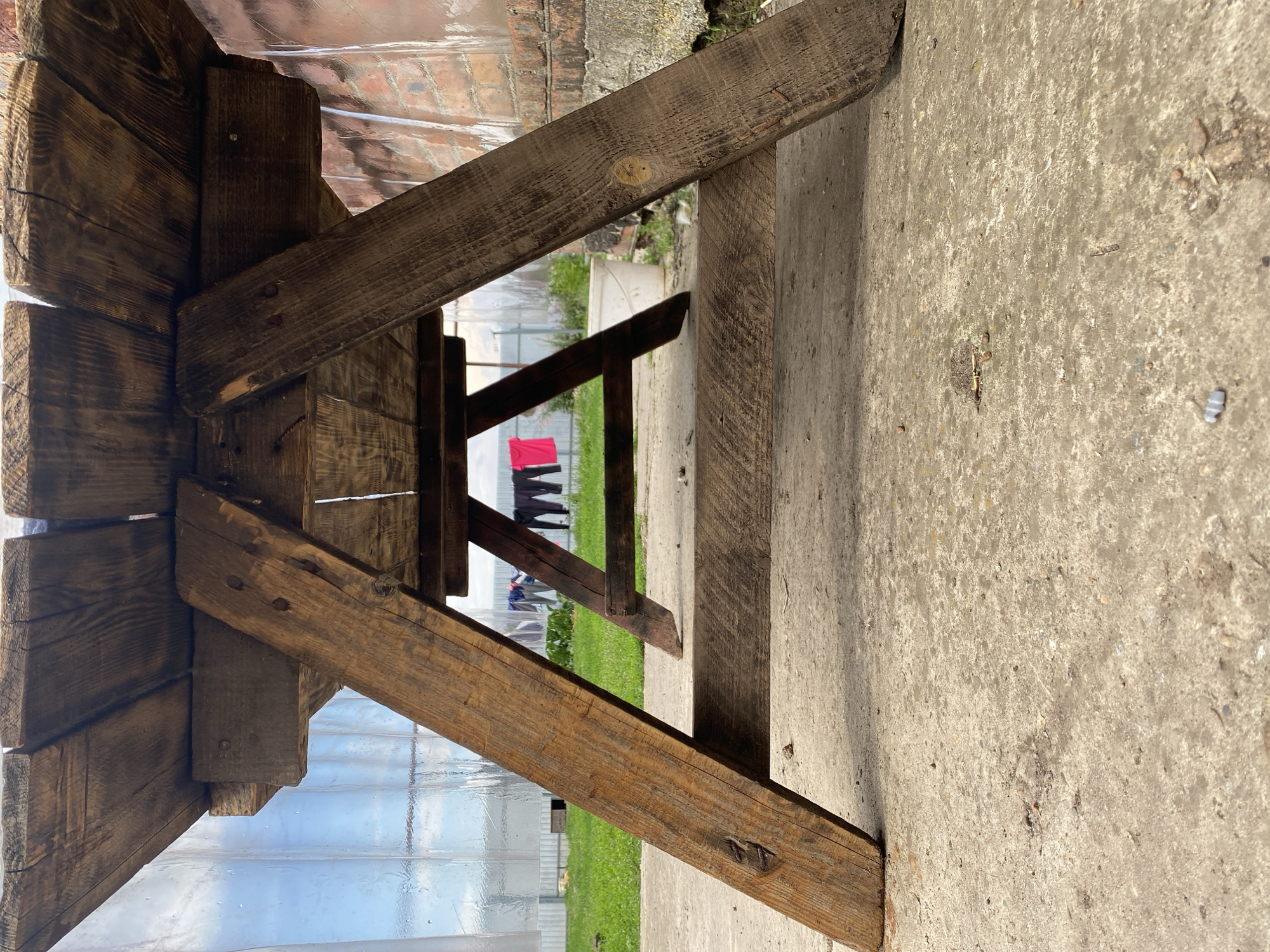The image shows the underside of a rustic, wooden picnic table, viewed from a low angle. The table is constructed from rough-hewn planks, with visible nails and grain. The legs of the table are made of thick, angled wooden beams. In the background, a green lawn is visible, behind which is a translucent plastic structure, possibly a greenhouse or covered area. Further in the distance, clothing is hanging on a clothesline, including a bright pink shirt and dark pants. A white bucket is also visible on the lawn. The ground in the foreground is a concrete surface, with scattered debris. The overall scene suggests an outdoor setting, likely in a rural or semi-rural area. While the specific location in Kisliakovskaya, Russia is mentioned, there are no visual cues in the image to confirm this location. There are no people or interactions depicted in the image.