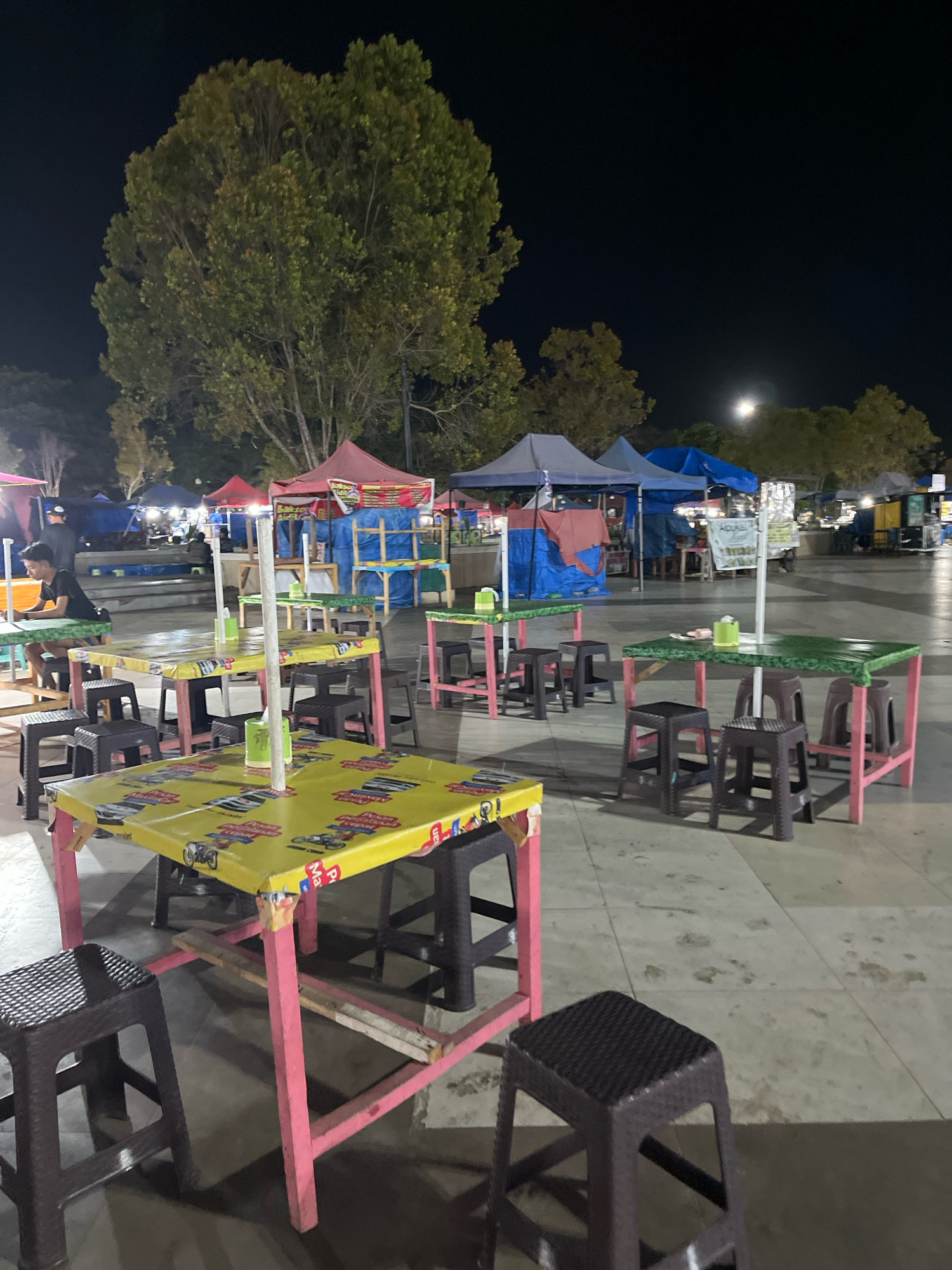 The image depicts an outdoor seating area at night, likely at a food stall or market in Kendari, Indonesia. Several tables with plastic tablecloths and accompanying stools are arranged on a paved surface. Some tables are covered in yellow plastic with colorful graphics, while others have green tops. The area is illuminated by artificial lighting, casting shadows. In the background, there are tents and structures that appear to be food stalls, some with blue and red tarpaulins. A few people are visible, including a young man sitting at a table and other figures in the distance, possibly vendors or customers. Trees with dense foliage are visible in the background, and the sky is dark, suggesting it is nighttime. The overall scene conveys a casual, open-air dining or gathering environment.