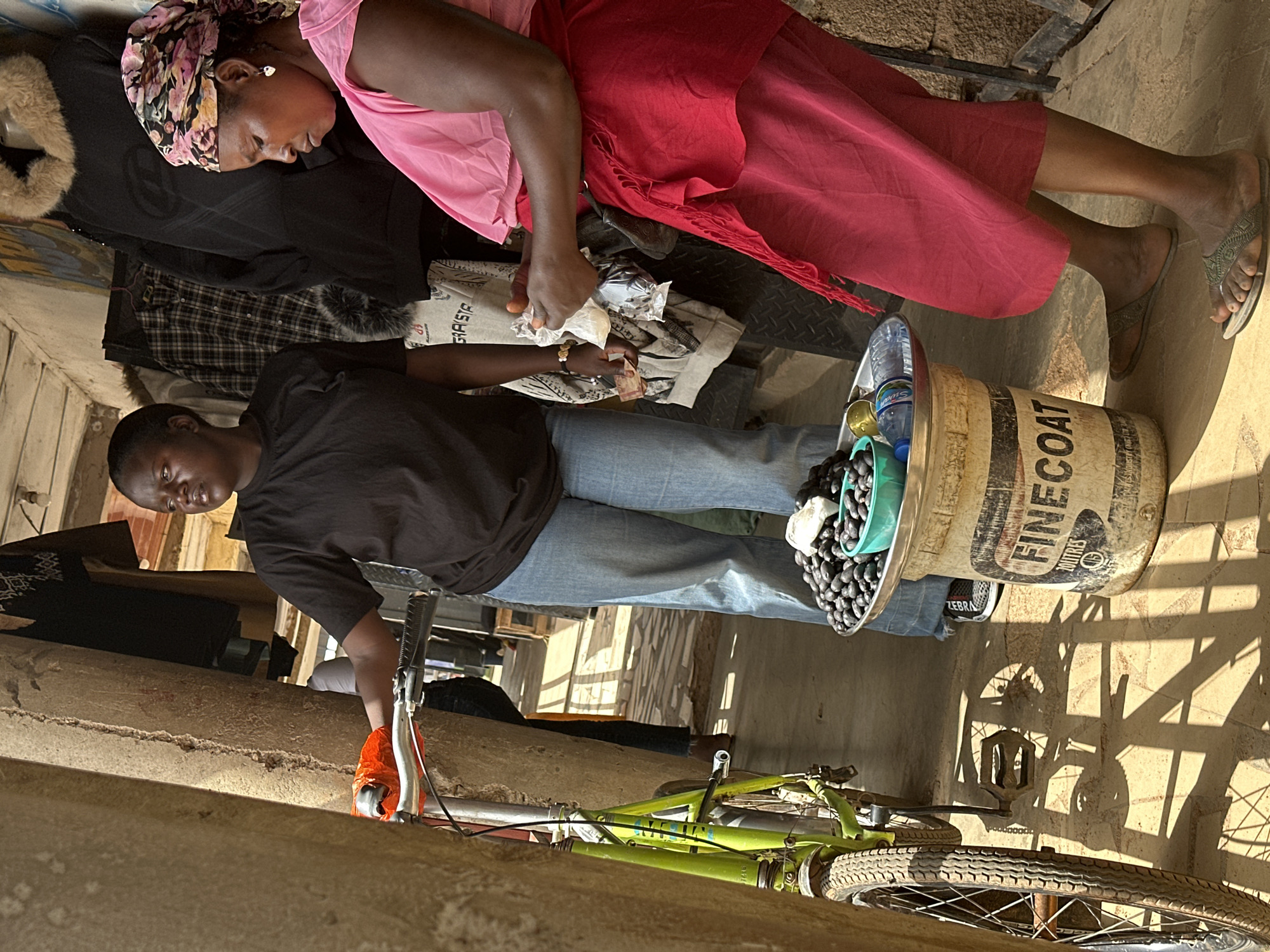 The image depicts a scene in Bukuru, Nigeria, where two individuals are interacting in an outdoor, possibly market or roadside, setting. In the foreground, a person wearing a black t-shirt and blue jeans is partially visible, leaning over a light green bicycle. They are wearing an orange glove on their left hand. A metal bucket, labeled "FINECOAT," is positioned to the right, filled with dark, round objects, possibly olives or berries, and water bottles. The bicycle is lying on its side.
In the background, another person is seated, wearing a bright pink top and a red wrap. They have a floral-patterned headscarf and are holding some items in their hands, appearing to be engaged in a transaction or exchange. The lighting suggests it is daytime, with strong shadows cast across the ground, indicating bright sunlight. The overall atmosphere seems informal and practical, possibly a moment of rest or commerce. Visible text on the bucket includes "FINECOAT" and "ZEBRA." The presence of the bicycle and the bucket suggests everyday life activities.
